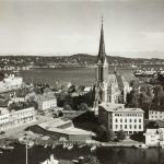 Arendal i 1948. Svart-hvitt foto av deler av Arendal sett ovenfra. Midt i bildet ruver Trefoldighetskirken med høyt, smalt spir og kors på toppen. Rundt kirken ligger tett bebygde kvartaler med lave tre- og murhus. I forgrunnen er det sjø med flere småbåter fortøyd langs brygger og kaier. I bakgrunnen strekker sjøen seg innover, med skogkledde åser og spredt bebyggelse på den andre siden av vannet.
