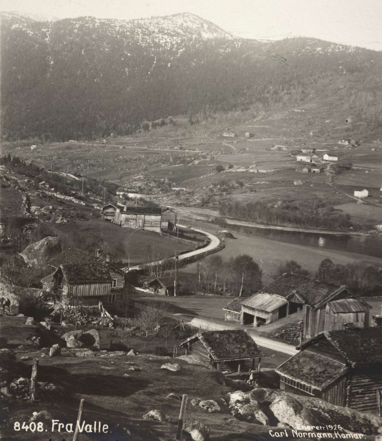 Dette er et svart-hvitt fotografi fra 1926, tatt av Carl Normann, som viser et naturskjønt landskap fra Valle i Norge. Bildet fanger en dal med en elv som slynger seg gjennom landskapet, omgitt av fjell med snødekte topper i bakgrunnen. Foran i bildet sees flere små tømmerhus med torvtak, typiske for norske gårdsbruk i denne perioden. Noen av husene har vedstabler inntil seg. En grusvei snor seg gjennom dalen, forbi et større, mer moderne hus med hvite vinduskarmer lenger ned i bildet. På den andre siden av elven ligger flere gårder spredt utover de skrånende jordene. Terrenget er kupert med steiner og små trær spredt rundt i landskapet. Bildet gir en stemningsfull fremstilling av et norsk bygdelandskap tidlig på 1900-tallet, preget av tradisjonelle byggeskikker og en vakker, men røff natur.