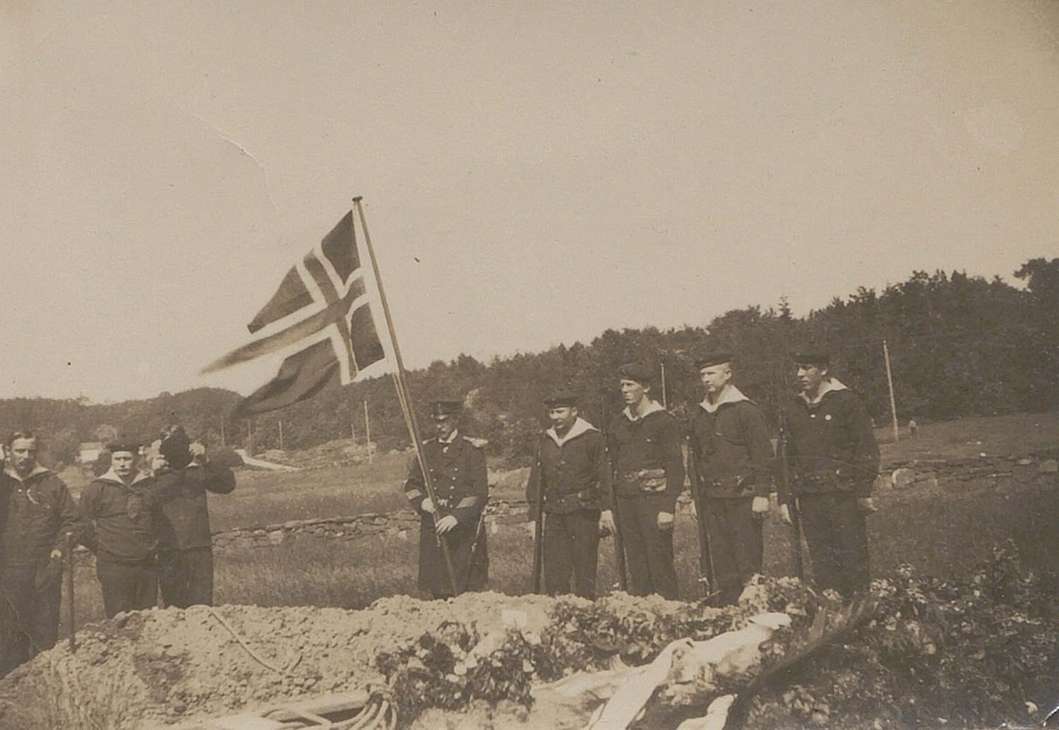 Bildet viser en begravelse på Høvåg kirkegård. I forgrunnen ligger en grav dekket med blomster og kranser. Rundt graven står en gruppe mennesker, blant dem norske marinefolk i uniform. Én person holder et norsk flagg med kors. Personene står stille og samlet, vendt mot graven. I bakgrunnen sees et naturlandskap med trær og åser, som gir en rolig ramme rundt seremonien. Bildet er tatt utendørs i dagslys. Begravelsen gjelder en ukjent tysk marinegast, og fant sted under første verdenskrig.