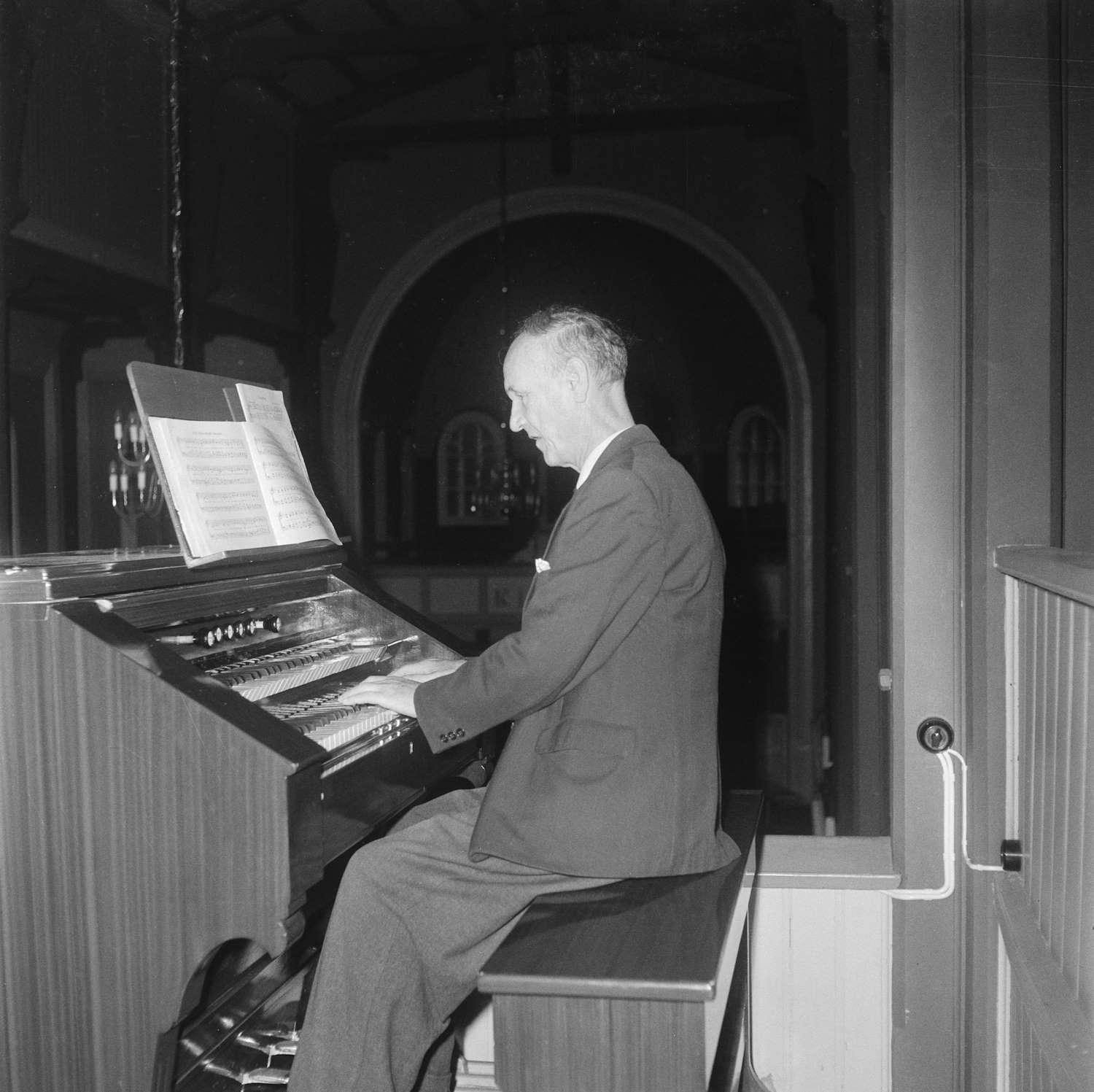 Bildet viser Georg Hørsdal i ferd med å spille orgel i en kirke. Han sitter på en benk foran det store orgelverket, kledd i dress, med hendene plassert på tangentene. Et notestativ med noteark står foran ham, og han er konsentrert om musikken. I bakgrunnen skimtes kirkens interiør med buede vinduer og en dempet, varm belysning som gir rommet en høytidelig og rolig atmosfære.