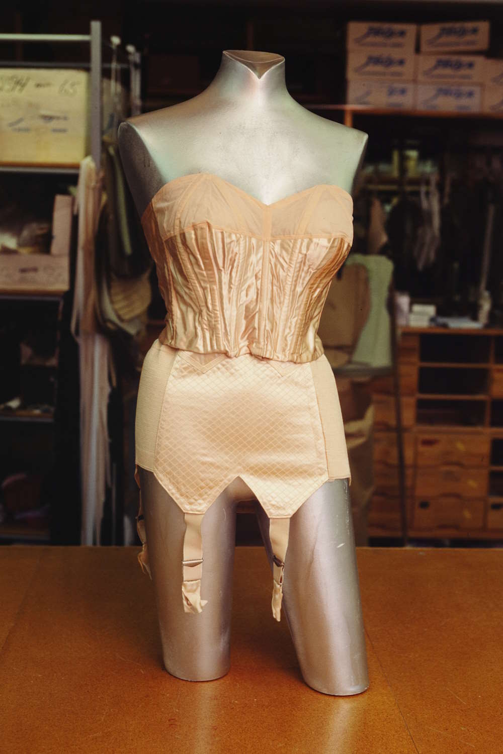 Korsett og hofteholder fra 1920-tallet utstilt hos Frøja konfektions AS, 18.03.1995, Fædrelandsvennen. Foto: Trygve SkramstadBildet viser en sølvfarget mannequin som står plassert på et bord. Mannequinen er kledd i et vintage korsett og hofteholder i en lys beige farge. Korsettet har tydelige vertikale sømmer og former kroppen i en klassisk timeglassfigur. Hofteholderen har fire stropper som henger ned foran. I bakgrunnen er det hyller med esker og ulike tekstiler, noe som gir inntrykk av at bildet er tatt i et lager eller verksted for klær eller kostymer.