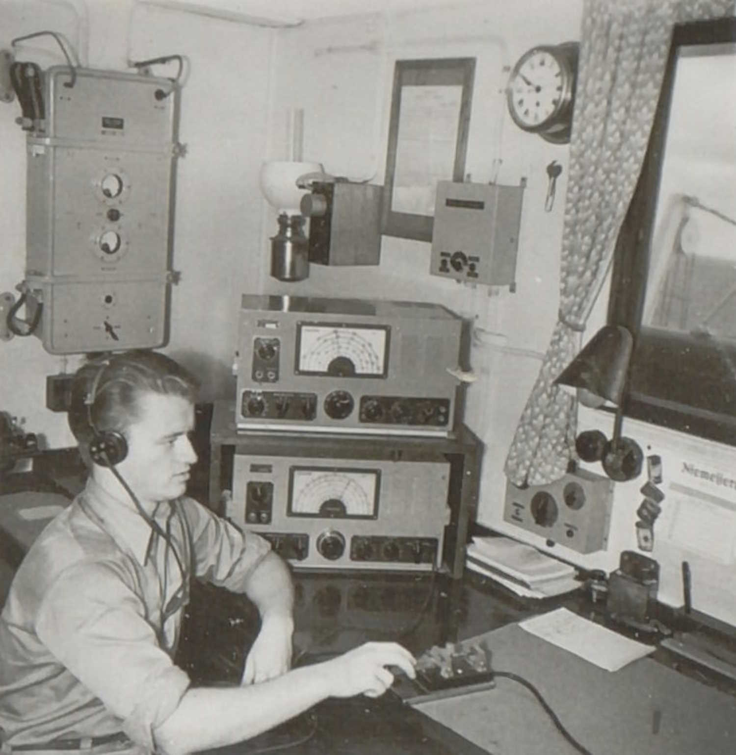 Bildet viser Ingebrigt Olsen i arbeid i radiorommet om bord i motortankeren Vardaas i 1951. Han sitter ved et skrivebord med hodetelefoner på, konsentrert om å betjene radioutstyret foran seg. På bordet ligger flere radioapparater med måleinstrumenter, en mikrofon og noen papirer, som antyder en aktiv kommunikasjonssituasjon. Veggene rundt er tett utstyrt med teknisk utstyr, inkludert en veggmontert klokke og en lampe som gir arbeidslys. Et vindu med gardiner gir innsyn til skipets dekk og slipper dagslys inn i rommet, noe som skaper en balanse mellom det tekniske og det hverdagslige. Bildet formidler en følelse av presisjon og ansvar, og gir et autentisk innblikk i sjømannens arbeidsmiljø på 1950-tallet.