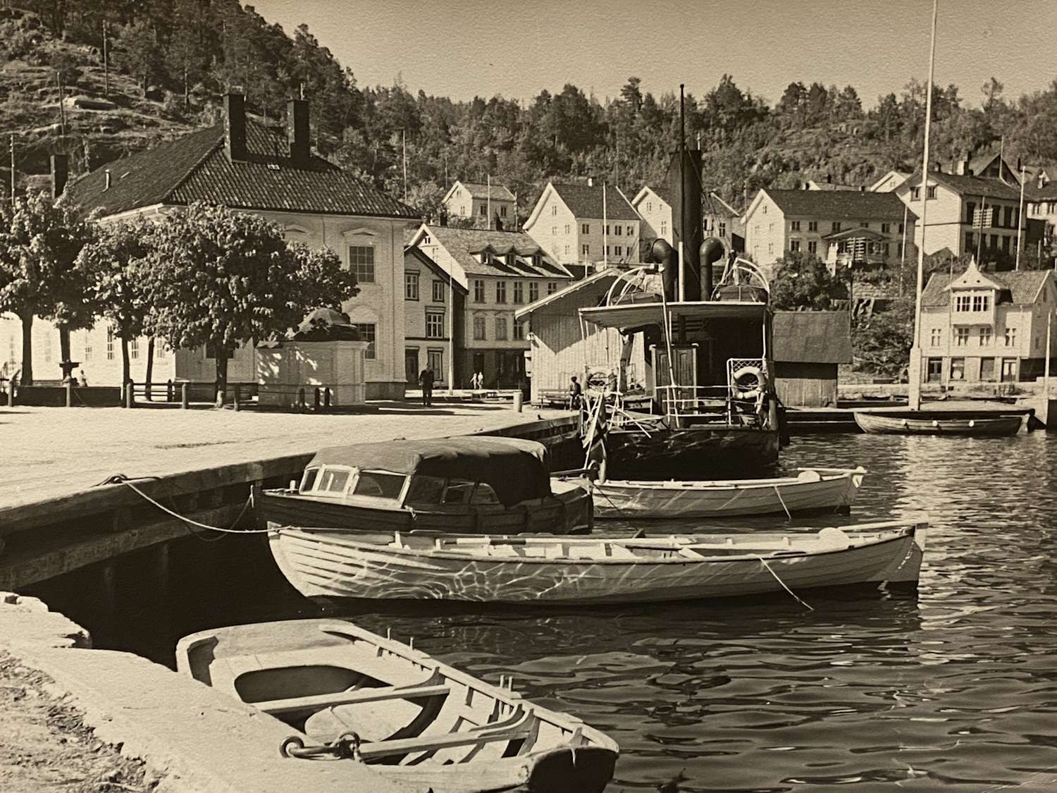 Tvedestrand 1951. Foto: Birger Dannevig. Bildet viser en rolig og idyllisk havn. I forgrunnen ligger tre båter fortøyd ved bryggen. Den nærmeste er en liten robåt, den midterste en motorbåt med overbygg over førerplassen, og den tredje en større båt med et rørformet utløp på dekket. Langs bryggen vokser trær som gir skygge til området. I bakgrunnen strekker det seg flere hvitmalte hus og bygninger langs vannkanten og oppover en skogkledd åsside. Hele scenen har et nostalgisk preg, og bildet er tatt i svart-hvitt eller sepia-toner, noe som gir det et historisk uttrykk.