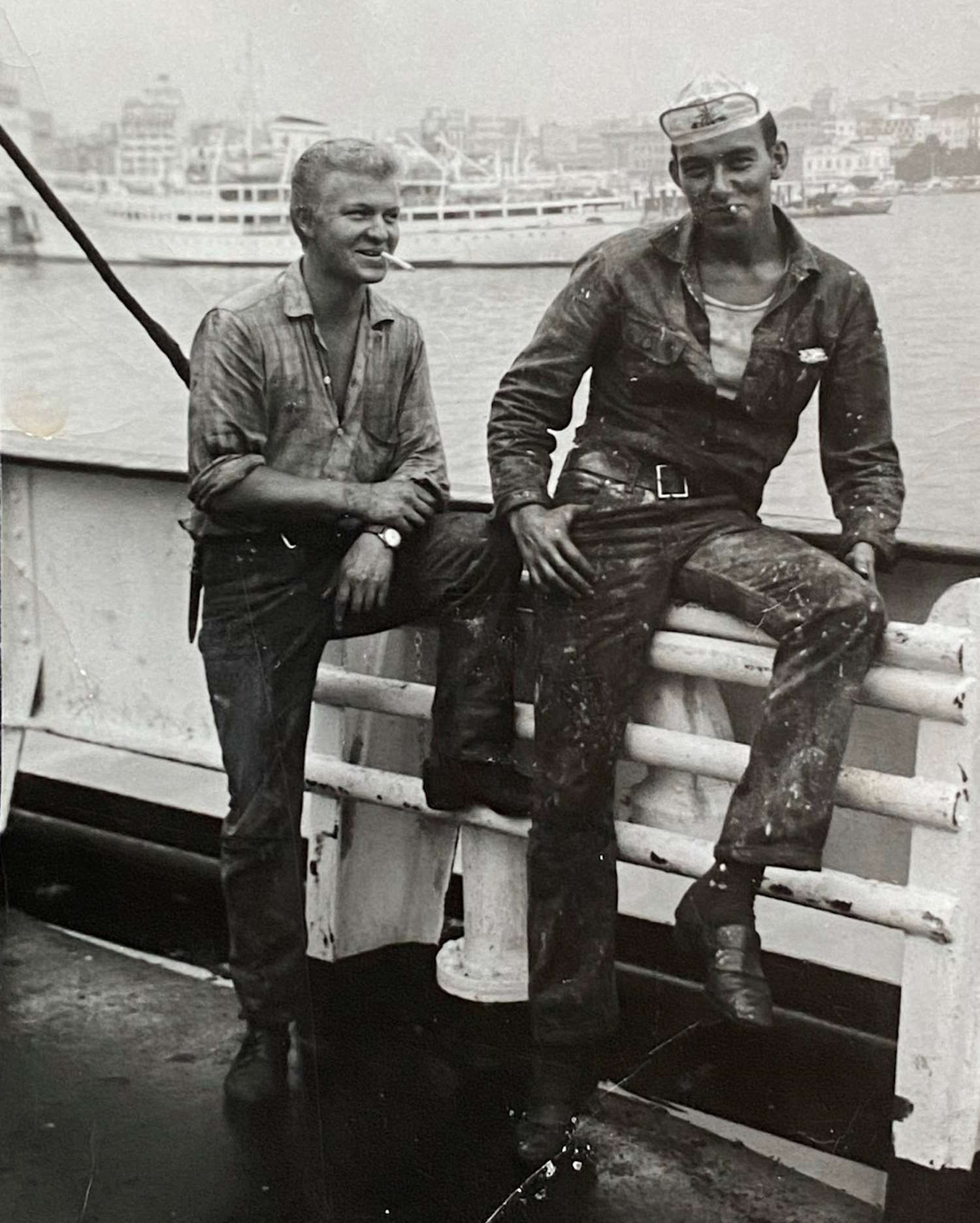 Dag Albert Witzøe Molaug og en skipskamerat om bord i "Concordia Sky" Liggende i Pireus 1962. Personene står på dekk nær en rekkverk-konstruksjon. Den ene personen står oppreist med armene avslappet, mens den andre sitter uformelt på rekkverket med det ene beinet hengende ned og det andre støttet mot en metallstang. Begge har arbeidsklær som ser godt brukt ut, med flekker og slitasje – typisk for mannskap som jobber på skip. Den som sitter har en lue eller caps på hodet. Underlaget på dekket ser mørkt og vått ut, som om det nylig har regnet eller blitt spylt. I bakgrunnen skimtes havneområdet med flere bygninger og en rekke mindre fartøy, blant annet en stor hvit båt med flere vinduer og overbygg. Det er en disig eller overskyet dag, noe som gir bildet et grålig preg.