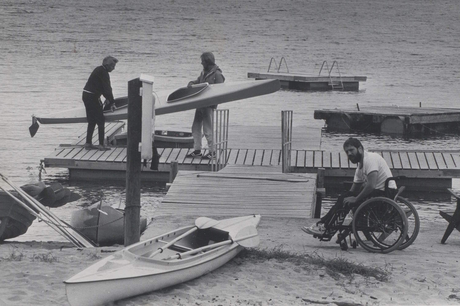 Svart-hvitt-bilde fra Hoveleiren på Tromøya en gang på 1980-tallet. Bildet viser en brygge ved sjøen, brukt under en årlig treningsleir for personer med fysiske funksjonsnedsettelser. I forgrunnen ligger en kajakk på sandstranden. Til høyre sitter en person i rullestol på sanden, vendt mot brygga. Midt i bildet er en tre-brygge med rampe ned mot vannet. To personer står på brygga og holder en kajakk mellom seg. I bakgrunnen ligger to flytebrygger ute på vannet. Vannet er rolig, og det er ingen synlige bølger.