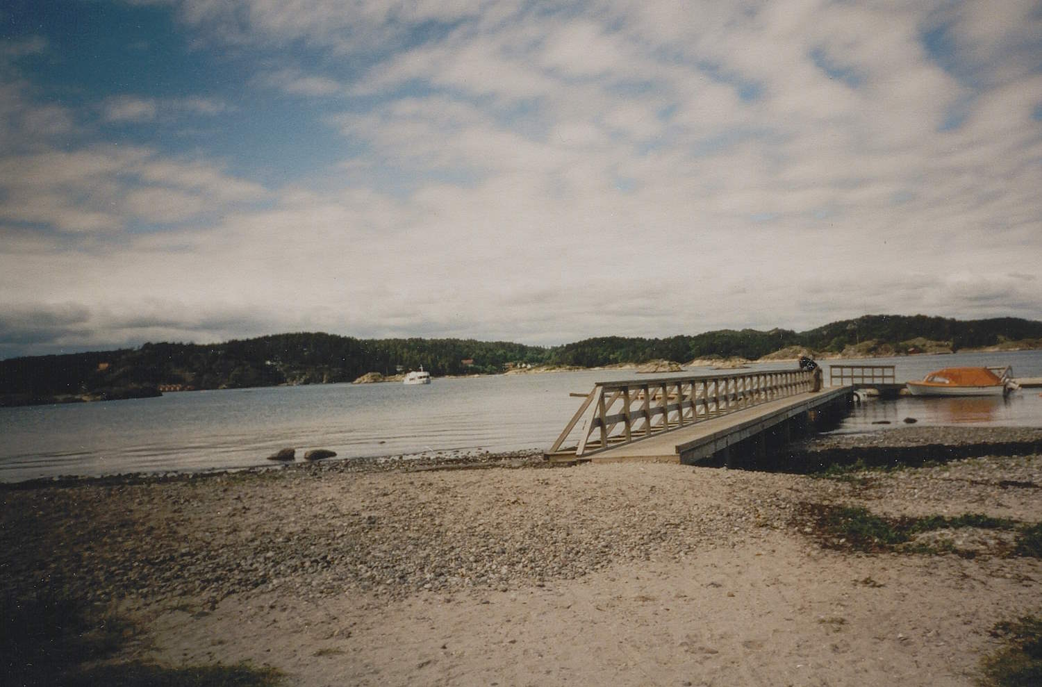 Fargefoto fra Hove på Tromøya, tatt i forbindelse med en treningsleir for personer med fysiske funksjonsnedsettelser. Bildet viser en strand med småstein og sand i forgrunnen. Midt i bildet går en lang trebro med rekkverk ut i sjøen, og ender i en liten brygge. Til høyre for brygga ligger en flåte med en oransje gjenstand oppå, trolig en båt eller kano. I bakgrunnen sees rolig sjø med noen småbåter og lave, skogkledde øyer under en himmel med spredte skyer.