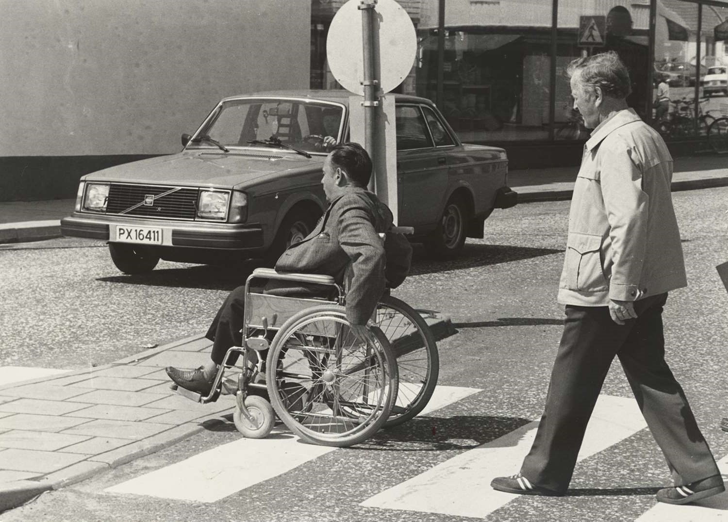 Rullestolbruker på vei over gangfelt. Fædrelandsvennens fotoarkiv. Fotograf: Jan A. Johannesen. Svart-hvitt foto fra 1970–80-tallet i et bymiljø. En person i rullestol og en annen person krysser et gangfelt, mens en bil står stille foran dem. En stolpe med trafikkskilt er synlig midt i bildet.