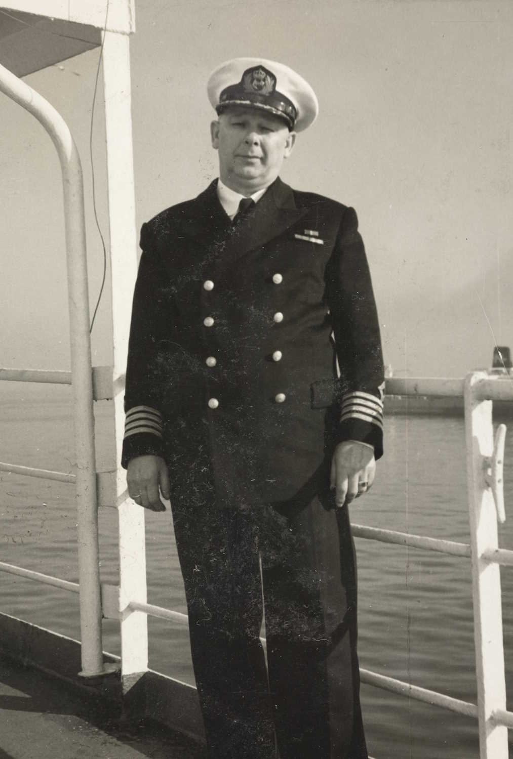 Bildet viser skipsfører Finn Abrahamsen stående på dekk av et fartøy. Han er kledd i en formell uniform som signaliserer høy rang til sjøs. Uniformen består av en mørk dobbeltspent jakke med seks blanke knapper i to vertikale rekker. På skuldrene er det epåletter, og på ermene sees flere brede striper, typisk rangtegn for en skipsfører eller kaptein. Finn har en hvit uniformslue med et tydelig emblem foran, som indikerer maritim tilknytning. Bakgrunnen viser rekkverket på skipet og et rolig havområde, med vann som strekker seg utover. Det er ingen synlige bølger, noe som gir inntrykk av stille vær. Bildet er i svart-hvitt, noe som tyder på at det er tatt i en tidligere tidsepoke, sannsynligvis midten av 1900-tallet. Teksturene på uniformen og rekkverket er tydelige, og lyset virker naturlig, som om bildet er tatt utendørs i dagslys