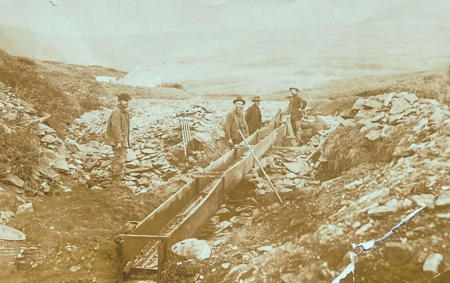 Bildet viser en historisk scene fra gullgraving i Alaska mot slutten av 1800-tallet. Fire personer står ute i et steinete landskap omgitt av lave gresskledde skråninger. Midt i bildet ligger en lang renne av tre, kjent som en «sluice box», som ble brukt til å vaske gullholdig grus med vann slik at det tunge gullet sank til bunnen. Personene er kledd i tidstypiske arbeidsklær med hatter og støvler, og de holder spader og andre redskaper. Rundt rennen ligger stein og jord, tydelig tegn på manuelt arbeid. I bakgrunnen skimtes et åpent, kupert landskap uten trær, og himmelen virker lys og overskyet. Bildet har et sepiafarget preg, typisk for fotografier fra denne perioden.
