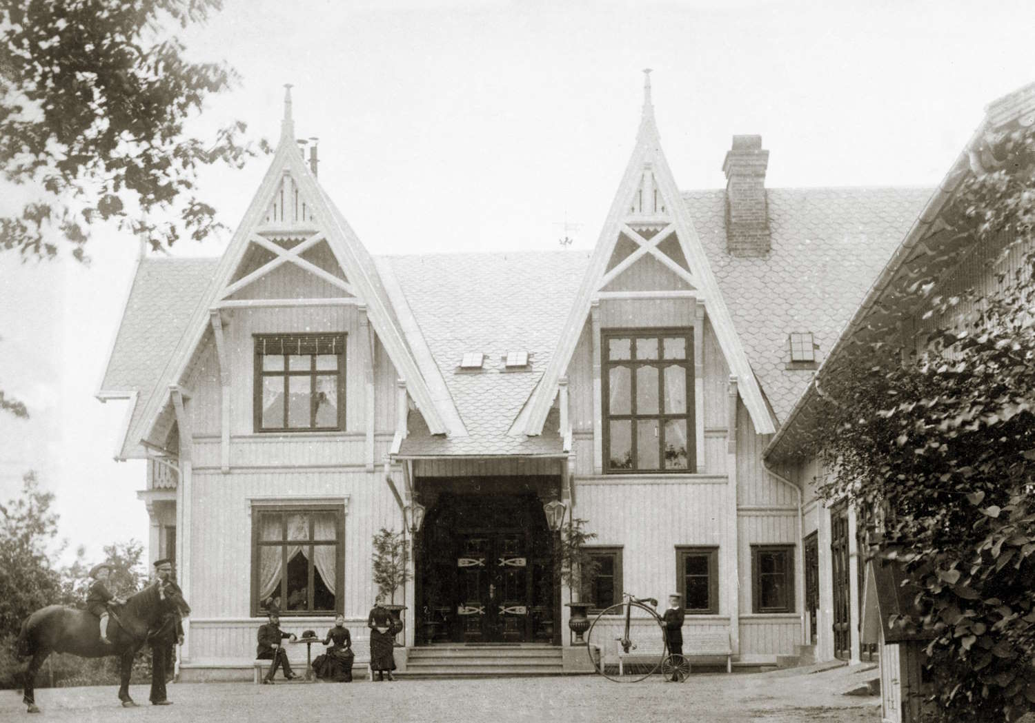 Axelhus med familien Herlofson i 1884. Foto: Per Adolf Thoren. Bildet viser en stor, herskapelig trevilla i sveitserstil med markante, spisse gavler og dekorative detaljer. Huset har to høye, symmetriske gavlpartier med store vinduer og et inngangsparti i midten med trapp og overbygg. Taket er tekket med skifer, og en stor murpipe er synlig til høyre. Foran huset står flere personer: to sitter på trappen, én står ved siden av, og en annen holder et høyt hjul – trolig en sykkel av eldre type (velosiped). Til venstre i bildet står en person med en hest. Til høyre skimtes en sidebygning med klatreplanter langs veggen. Bildet er i sort-hvitt.
