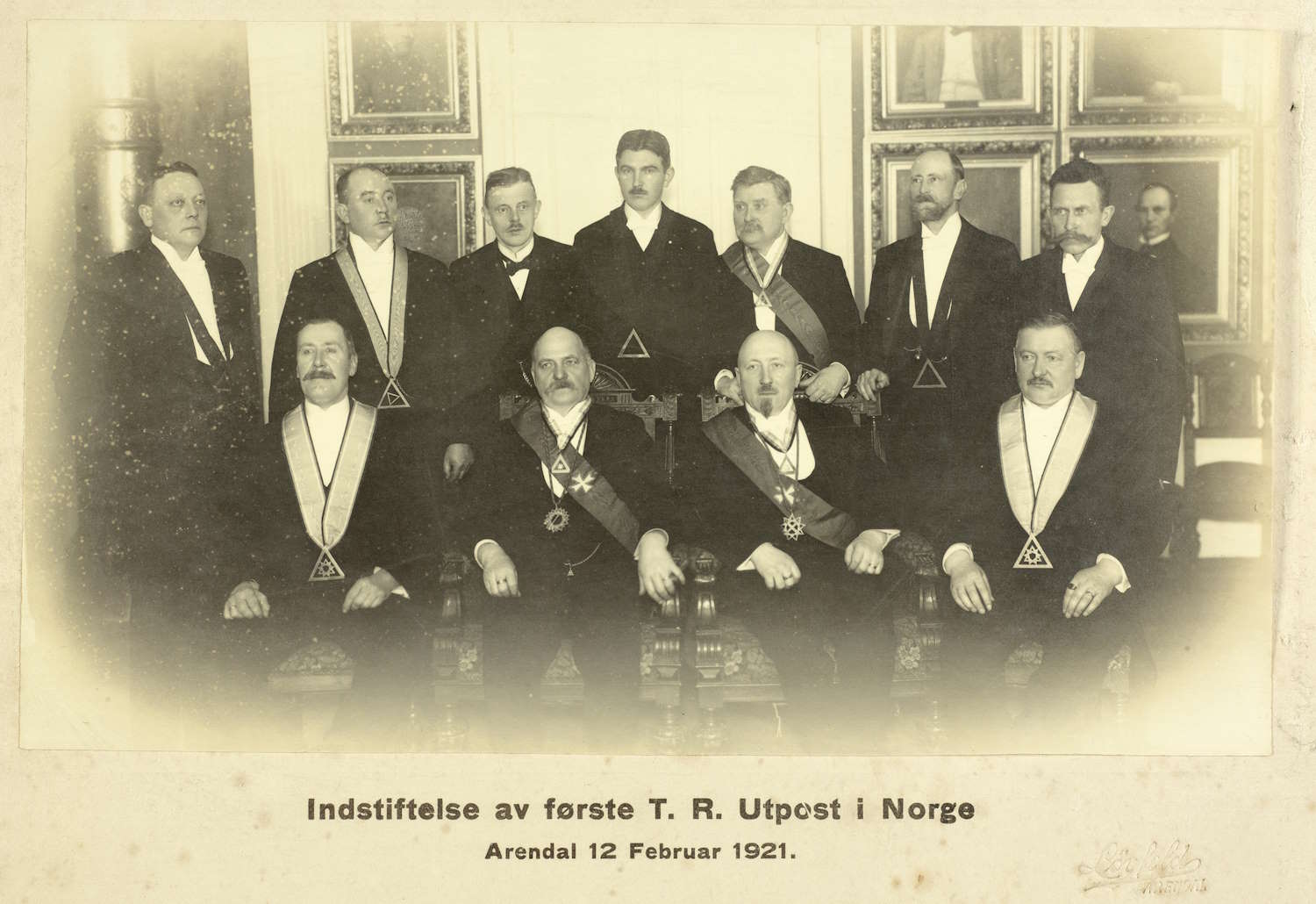 Innstiftelse av første Tempelridder utpost i Norge. Arendal 12.02.1921. Foto: Magnus Løvfold. Det er elleve personer kledd i mørke, høytidelige dresser med hvite skjorter og slips. Flere av dem bærer seremonielle bånd og medaljer, og noen har trekantformede symboler hengende foran seg. Personene sitter og står i to rekker – fem i front og seks bak. Bakgrunnen består av en elegant, klassisk interiørstil med store, innrammede malerier eller dekorative paneler på veggen. Bildet har en sepia-tone som gir det et gammelt, tidstypisk preg. Nederst på bildet er det en tekst som lyder: «Indstiftelse av første T. R. Utpost i Norge Arendal 12 Februar 1921.»