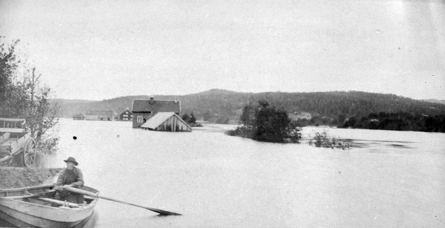 Messel i Froland under flommen i 1892. Sort-hvitt fotografi som viser en stor flom. Vann dekker nesten hele landskapet, og bare hustakene på noen bygninger stikker opp av vannet i midten av bildet. I forgrunnen, til venstre, sitter en person i en robåt med en åre, nær en trerot eller busk som stikker opp av vannet. Ved elvebredden står en vognhjul delvis synlig. I bakgrunnen er det skogkledde åser og flere hus som ser delvis oversvømt ut. Bildet gir et sterkt inntrykk av ødeleggelse og naturkrefter under flommen.