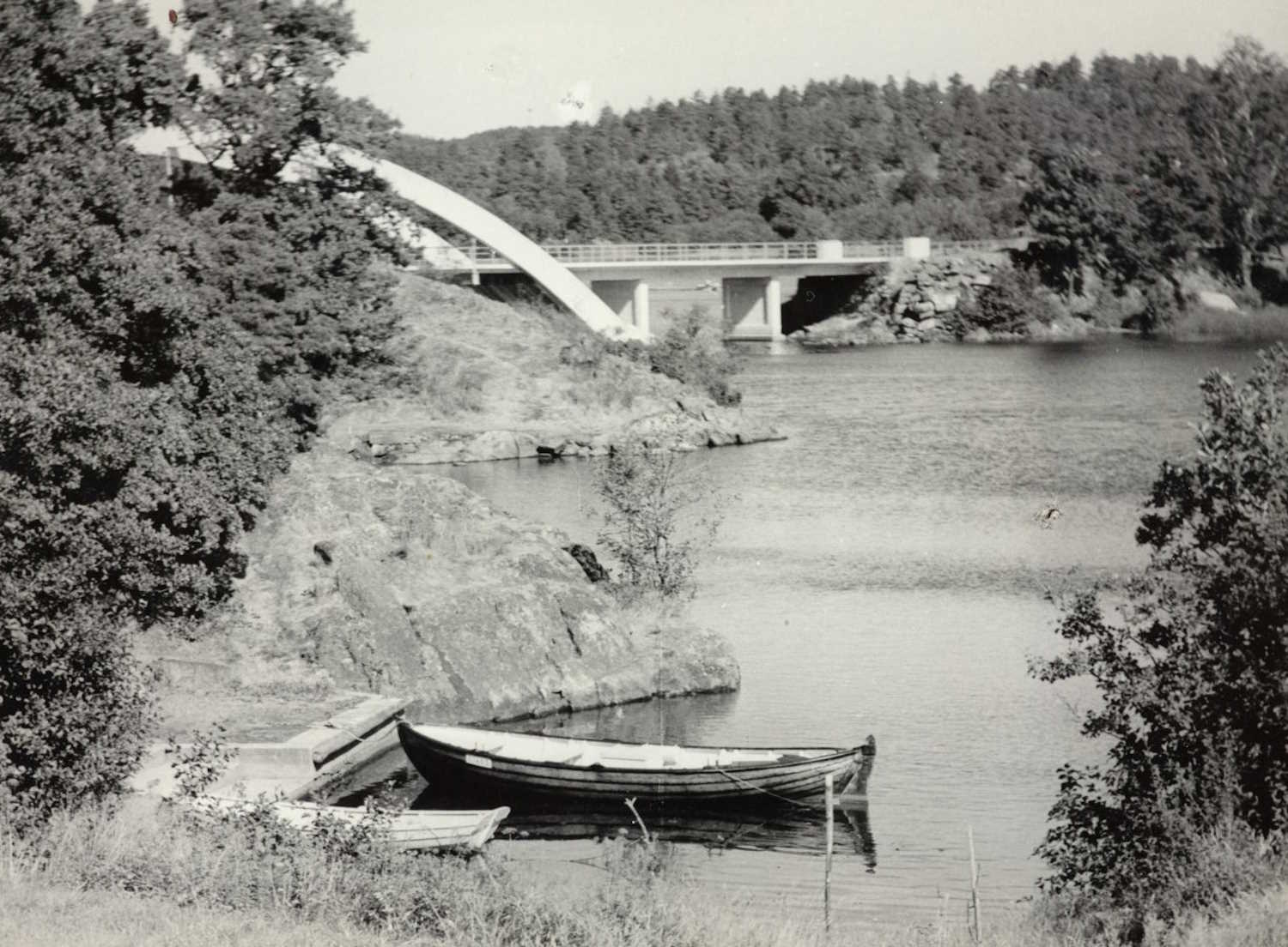 Parti av Vippa bro rundt 1960. Sort-hvitt fotografi som viser et elvelandskap med tett vegetasjon på begge sider. Til venstre står et stort tre med kraftige greiner, og til høyre er det busker og mindre trær. I bakgrunnen skimtes en bred elv som renner rolig, deler av Vippa bro, og skogkledde åser. I forgrunnen ligger to robåter av tre fortøyd til en liten brygge. Bildet gir et inntrykk av et stille, naturlig område ved vannen.