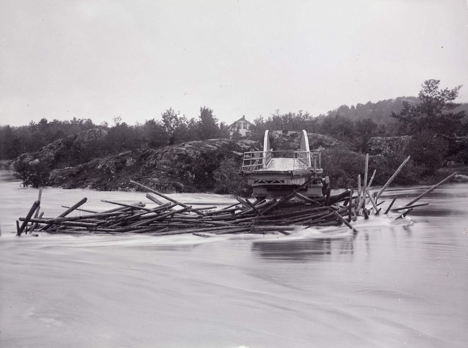 Vippa bro ødelagt av flommen i 1892. Foto. Frantz Leopold Stockinger. Sort-hvitt fotografi som viser Nidelva med sterk vannføring. Midt i bildet står restene av Vippa bro - en ødelagt brokonstruksjon – en liten bueformet stålkonstruksjon med rekkverk – som har blitt revet løs og ligger fastklemt mot en stor samling tømmerstokker i vannet. I bakgrunnen er det skogkledde åser og en enslig bygning på en høyde. Vannet i forgrunnen er urolig, og bildet gir et inntrykk av flomskader og naturkrefter.