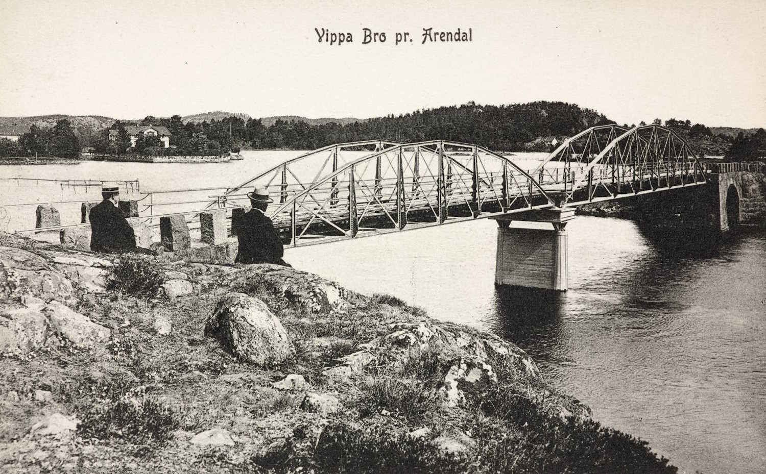 Sort-hvitt fotografi som viser en stor metallbro med buer og gitterkonstruksjon som strekker seg over en bred elv. Broen har solide steinmurer på hver side og en betongpilar midt i vannet som støtter konstruksjonen. I forgrunnen er det et steinete område med gress og noen store steiner, der to personer sitter og ser mot broen. Bak broen ligger skogkledde åser og et par bygninger ved vannkanten. Øverst i bildet står teksten: «Vippa Bro pr. Arendal». Bildet gir et inntrykk av ingeniørkunst og landskap tidlig på 1900-tallet.