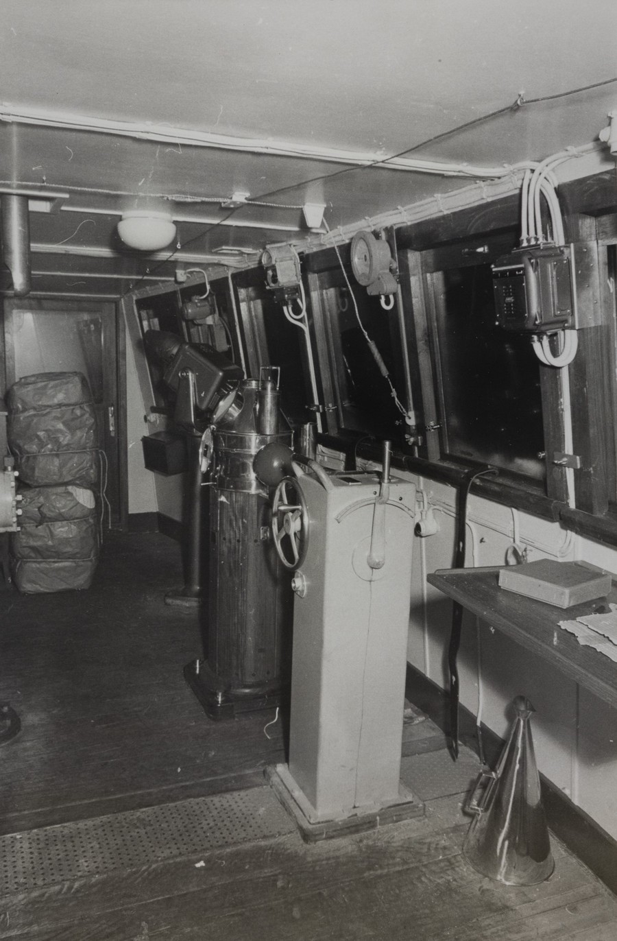 Bildet er et svart‑hvitt fotografi som viser broa på lasteskipet M/S "Arendal" i 1955. Rommet er avlangt og smalt, med treverk på gulvet og store vinduer langs høyre side. Utenfor vinduene er det mørkt, slik at ingenting av omgivelsene er synlig. Midt i rommet står navigasjonsinstrumenter fra 1950‑tallet. Nærmest kameraet er en høy, firkantet styrekonsoll med et ratt øverst. Bak denne står et større, rundt instrumenthus i metall, med kompass øverst og flere håndtak og brytere. Lenger bak i rommet skimtes flere apparater og mekaniske innretninger som brukes til styring og navigasjon på skip. Langs høyre vegg henger kabler og et firkantet, veggmontert instrument. Under vinduene går en smal hylle med blant annet en kasse og noen papirer. På gulvet til høyre står en stor, traktformet metallgjenstand, trolig et talerør eller en del av skipets kommunikasjonssystem. I bakgrunnen, mot venstre, ligger flere store, innpakkede kolli eller sekker stablet oppå hverandre. Belysningen i rommet er enkel, med et lavt tak og en rund kuppellampe. Stemningen i bildet er teknisk og funksjonell, som en arbeidsplass der alt på broa var manuelt og mekanisk betjent.