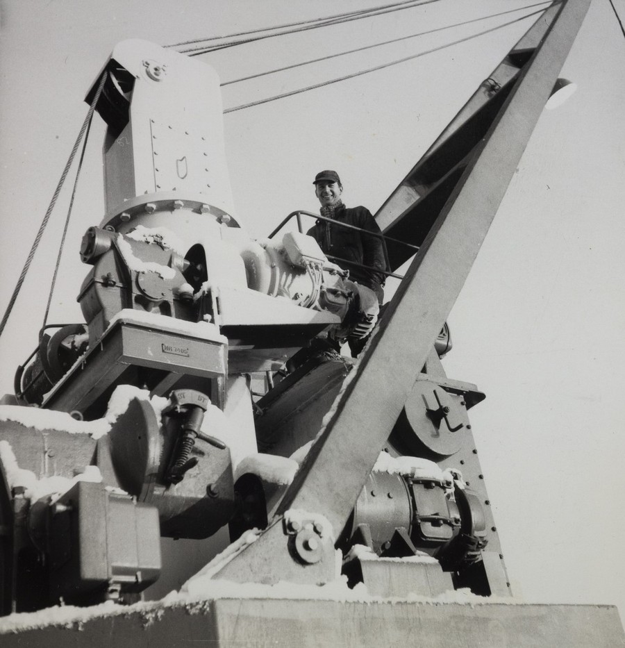 Bildet er et svart‑hvitt fotografi som viser en av dekkskranene om bord på lasteskipet M/S "Arendal". Kranen fyller store deler av motivet og framstår som en tung, mekanisk konstruksjon i metall med mange detaljer, bolter og ledd. Den står på et solid fundament og har en skråstilt bom som strekker seg opp mot høyre side av bildet. Flere vaiere går fra toppen av kranen og videre ut av rammen. I forgrunnen sees ulike tannhjul, motorhus og hydrauliske eller mekaniske komponenter dekket av et lett lag med rim eller snø, noe som antyder at bildet er tatt i kaldt vær. Overflater og kanter har en grov, industriell karakter som er typisk for midten av 1900‑tallets lasteutstyr. Bak på kranens plattform står en person i arbeidsantrekk, støttet mot et gelender. Kroppen er tydelig, men ansiktet er uten detaljer. Personen gir inntrykk av å være i ferd med å inspisere eller betjene kranen. Bakgrunnen er lys og ensfarget, uten synlige bygninger eller landskap, som gir motivet et klart fokus på kranens mekanikk og størrelse.