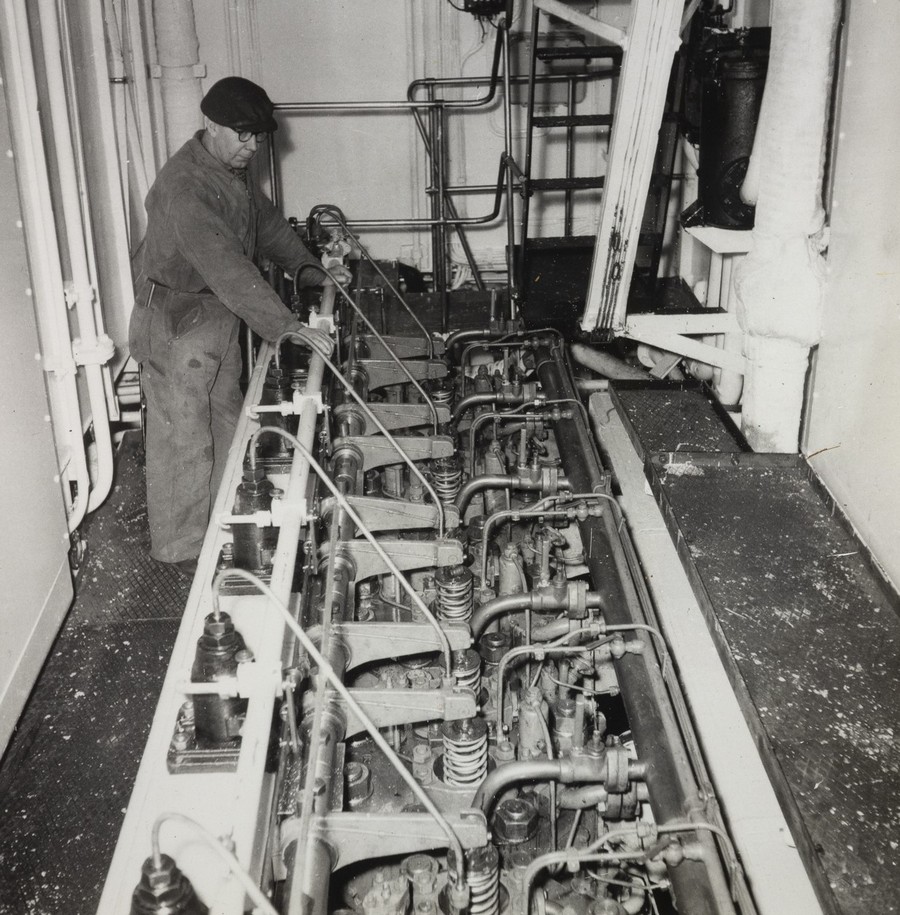 Bildet er et svart‑hvitt fotografi fra maskinrommet om bord på M/S "Arendal". Motivet viser en stor del av skipets motor installert nedsenket i en dyp motorbrønn. Motoren fyller hele den nedre delen av bildet, med mange synlige komponenter: ventiler, stenger, fester, rør og fjærer. Mekanikken fremstår tung, detaljert og robust, typisk for midten av 1900‑tallets skipsmotorer. Langs venstre side av motoren står en person i arbeidstøy. Personen holder begge hendene på en lang, horisontal stang som ser ut til å være en del av motorens betjenings- eller reguleringssystem. Arbeidsklærne består av en overall og lue, og kroppsholdningen tyder på at vedkommende er i ferd med å inspisere eller justere motoren. Rommet rundt motoren har et teknisk preg med rør, ventiler og metallflater langs veggene. Flere rørføringer er festet i mønstre på bakveggen, og på høyre side står en opphøyd metallplattform med sklisikker overflate. En stige fører opp til et nivå over maskinrommet, delvis synlig øverst i bildet. Totalt gir fotografiet et innblikk i et aktivt og arbeidskrevende maskinrom, med den store motoren som dominerende sentrum i en tett og funksjonell teknisk installasjon.