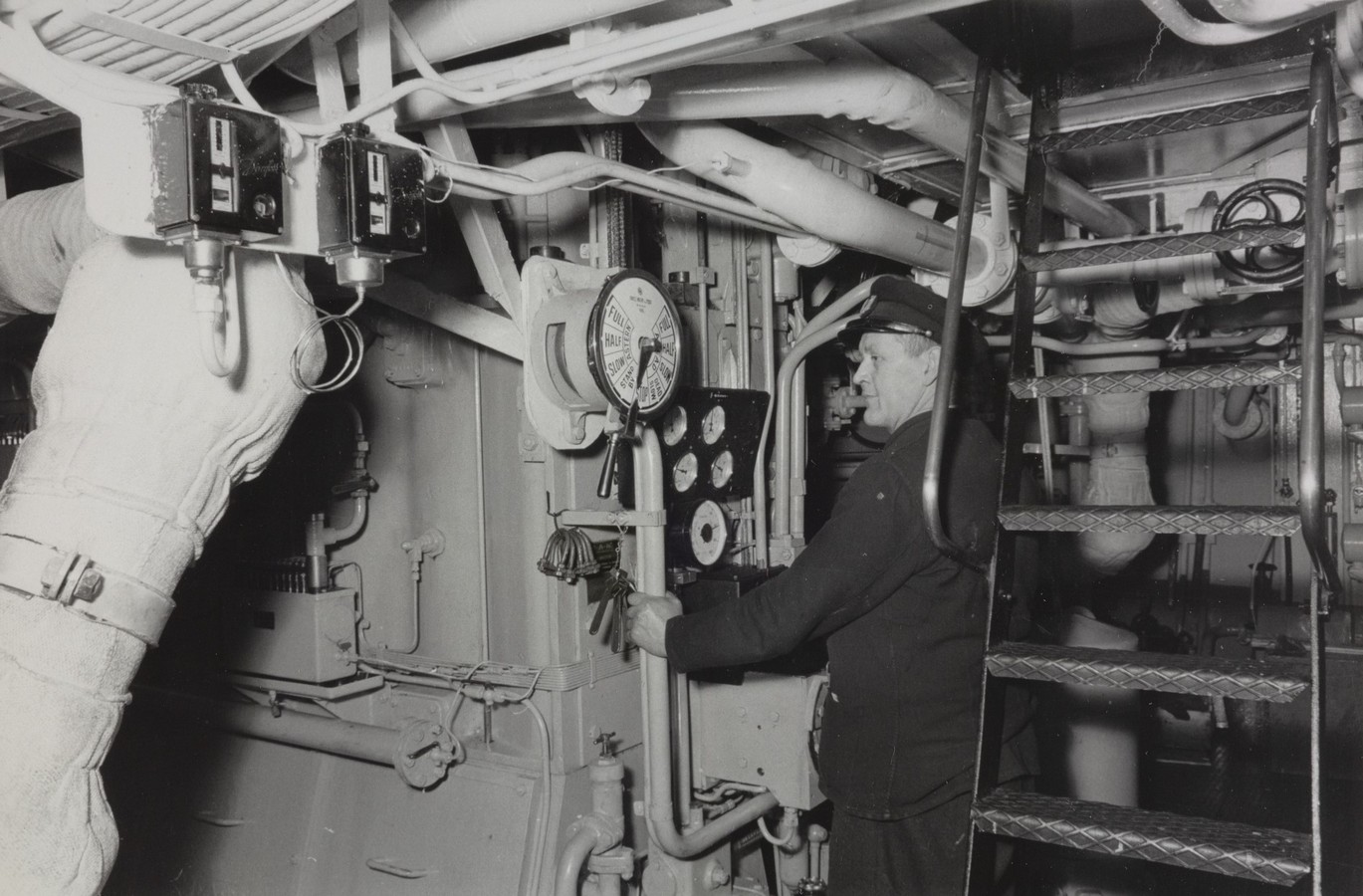 Bildet er et svart‑hvitt fotografi som viser en teknisk arbeidsstasjon inne i maskinrommet på M/S "Arendal". Rommet er tett pakket med rør, instrumenter og maskineri. Det domineres av lyse, kraftige rørledninger som går i ulike retninger over hodehøyde, festet til tak og vegger. I midten av motivet står en person i arbeidstøy foran et instrumentpanel. Panelet er montert på veggen og består av flere runde målere med visere, pluss brytere og hendler. Over panelet er det et stort, rundt måleinstrument med tallskive, festet til et loddrett stativ. Til høyre i bildet står en metalltrapp med gittertrinn som leder opp til et høyere nivå i maskinrommet. Under trappen og i bakgrunnen ses mer rørføring, ventiler og tekniske komponenter, som gir et inntrykk av et komplekst og kompakt maskinmiljø. Øverst til venstre er to små rektangulære apparater festet i en underside av en rørseksjon. Rørene rundt er isolert med stoff- eller fibermateriale, typisk for damp- eller varmeledninger fra perioden. Stemningen i bildet er industriell og funksjonell, med fokus på arbeid, drift og overvåking av skipets maskineri.