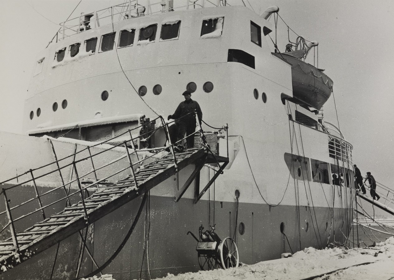 Bildet er et svart‑hvitt fotografi som viser M/S "Arendal" fortøyd ved kai vinterstid. Skipets side dominerer motivet, og det ligger et tykt lag snø og is både på kaien og på skipets skrog og overbygg. Fra skipet og ned mot kaien går en bred leider (gangvei) med rekkverk på begge sider. Den står i bratt vinkel og er snødekt. Midt på leideren står en person kledd i arbeidsantrekk, på vei ned. En kraftig fortøyningstrosse henger langs skutesiden. I forgrunnen, nede ved kaien, står et lite hjultrukket apparat – sannsynligvis en mobil vinsj, pumpe eller et annet arbeidsredskap – delvis omgitt av snø. Lenger akter på skipet, til høyre i bildet, klatrer flere personer opp en annen, smalere leider. Skipets overbygg ruver i bakgrunnen. Det har flere rekker av vinduer, runde koøyer og et livbåtstativ med en livbåt hengende på siden. Overbygget er også dekket av rim og snø, som gir hele motivet et kaldt, vinterlig preg.
