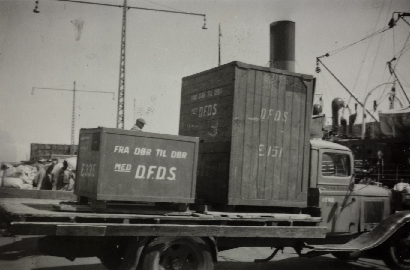 Bildet er et svart‑hvitt fotografi fra en havneområde i 1955. I forgrunnen står en lastebil med flatt lasteplan. På lasteplanet er det to store trekasser. Den største kassen står bakerst og er høy og rektangulær. Den mindre står foran den og er bredere og lavere. Begge kassene er merket med teksten “DFDS”, og den minste har i tillegg påskriften “FRA DØR TIL DØR MED DFDS”. Lastebilen er en eldre modell med runde hjulskjermer og en førerhytte med en enkel dør og frontrute. Noe av motorpanseret og høyre side av hytten er synlig. Et menneske står ved lasteplanet og ser ut til å arbeide med lasten. I bakgrunnen sees en travel havnescene: kraner, master, tauverk og deler av et skip med mannskap om bord. Til venstre i bildet ligger sekker og annet lastemateriale stablet på bakken. Høye, slanke stålkonstruksjoner med hengende lamper eller kroker rager opp mot himmelen. Stemningen i bildet er industriell og hektisk, preget av typisk havneaktivitet midt på 1900‑tallet.