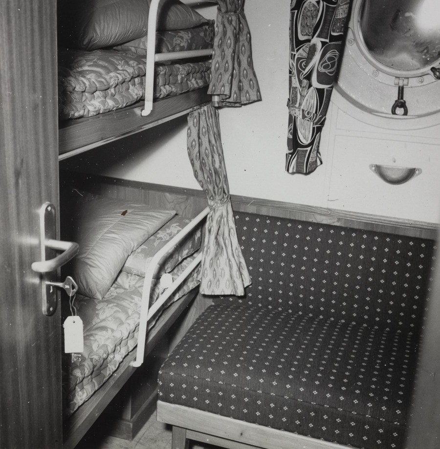 Bildet er et svart‑hvitt fotografi som viser en liten lugar om bord på M/S "Arendal". Interiøret er kompakt og funksjonelt, typisk for 1950‑tallets passasjer- og mannskapsrom. Til venstre i bildet står en køyeseng med to sengeplasser, én over den andre. Sengene har enkle madrasser og tykke tepper som ligger brettet. Øvre og nedre køye har hvite metallkanter og rekkverk som kan felles opp eller ned. En dørkarm og en delvis åpen dør er synlig helt til venstre. Til høyre for køyesengen står en polstret stol eller liten sofa med rygg og sete trukket i et tekstil med små, jevne mønstre. Over denne sitter et rundt skrogvindu (koøye) med metallramme, hengsler og lukkemekanisme. Rundt vinduet henger gardiner i et kraftig, mønstret stoff. En smal gardin henger også mellom køyesengen og veggen, muligens som enkel avskjerming for den nederste køyen. Rommet har ellers en rett og enkel veggkledning, og gulvet ser ut til å være dekket med linoleum eller annet robust materiale. Stemningen i bildet er maritim og praktisk — et lite, ryddig og typisk lugarrom fra et 1950‑tallsskip.