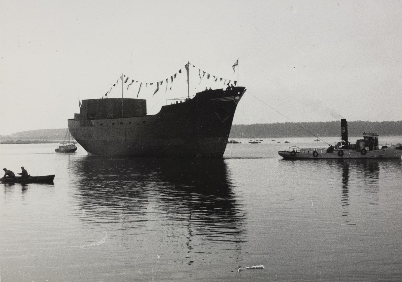 Bildet er et svart‑hvitt fotografi som viser skroget til M/S "Arendal" kort tid etter at det ble sjøsatt 10. november 1954. Skipet ligger høyt i vannet, uten overbygg og uten ferdig utrustning. Det fremstår som et stort, mørkt skrog med rette linjer og en markant baug. Langs skipets kant henger en rekke flagg og vimpler i snor, som er typisk ved sjøsetting. Skroget slepes eller manøvreres av en liten slepebåt på høyre side av bildet. Slepebåten er lav og kompakt, med en røykrørslignende skorstein. Vannet ligger nesten helt stille og speiler skipet. Til venstre i forgrunnen padler to personer i en liten robåt eller kano. Lenger bak i bildet sees flere småbåter og en svak horisontlinje med lavt land i bakgrunnen. Motivet gir et høytidsstemt inntrykk av en nybygget skute som nettopp har fått sitt første møte med havet.