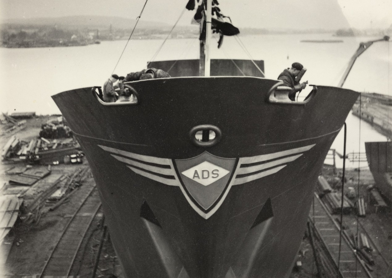 Bildet er et svart‑hvitt fotografi tatt like før sjøsettingen av M/S "Arendal" 10. november 1954. Motivet er sett forfra og nedenfra, slik at skipets baug fyller store deler av bildet. Baugen er nymalt og glatt, og på midten sitter rederimerket: et skjoldformet emblem med bokstavene ADS, flankert av to brede, stiliserte vinger. Langs øvre kant av baugen står flere personer og arbeider. De lener seg fram og holder i tau eller utstyr knyttet til sjøsettingsprosessen. Over dem henger flagg og vimpler festet til masten, som en del av markeringen. I bakgrunnen skimtes verftsområdet, med jernbanespor, trevirke og utstyr som ligger spredt på bakken rundt slippen. Lenger ute ses vannet, med rolige refleksjoner og et lavt landmassiv på den andre siden av fjorden. Silhuetten av større skip og lektere ligger langs horisonten. Bildeinntrykket er preget av forventning og aktivitet — et stort, nybygget skipskrog som står klar til å gli ut i vannet for første gang.