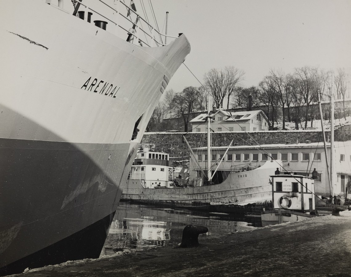 Bildet er et svart‑hvitt fotografi som viser M/S "Arendal" liggende til kai i Oslo i 1955, sammen med to andre fartøy. I forgrunnen, på venstre side, fyller baugen til M/S Arendal store deler av motivet. Skipssiden er malt i lyse og mørke felt, og navnet “ARENDAL” står tydelig skrevet på skroget nær baugen. I midten og til høyre i bildet ligger to mindre fartøy ved samme kaiområde. Ett av dem er en skøyte med navnet “TRIO” malt på skutesiden. Fartøyene ligger side om side langs kaifronten, med rigg, master og tauverk synlig. Et lite havnebygg og et skur står helt nede ved vannkanten. Lenger bak ser man en bratt skråning med trær som står uten løv, noe som tyder på vinter- eller tidlig vårsesong. På toppen av skråningen ligger flere bygninger, trolig bolighus eller mindre kontorbygg. Vannet i havnebassenget er stille og reflekterer skrogene. Delvis synlig på kaien står en mørk gjenstand som kan være en fortøyningspullert eller et stykke havneutstyr. Motivet gir et rolig inntrykk av en vanlig hverdag i havnen, med flere fartøy samlet langs kaiområdet i Oslo midt på 1950‑tallet.