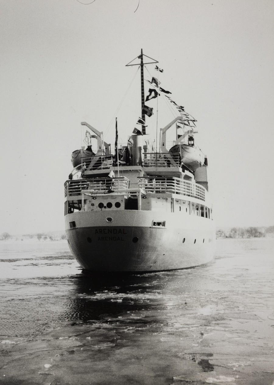 Bildet er et svart‑hvitt fotografi som viser akterenden på M/S "Arendal mens skipet beveger seg gjennom islagt farvann under jomfruturen i februar 1955. Skipet fyller nesten hele motivet og ligger midt i rammen. Akterskipet har en avrundet form, og navnet “ARENDAL” står malt i store bokstaver på hekken, med hjemstednavnet under. Langs de øvre dekkene sees rekker av rekkverk og ulike strukturer. To store livbåter er plassert på hver side av skipet i daviter. Midt på akterdekket står en mast med flere vaiere og en rekke vimpler og flagg som vaier i vinden, noe som gir inntrykk av en høytidelig anledning. Skipet beveger seg gjennom et vannspeil med issørpe og tynne isflak som er brutt opp av fremdriften. I bakgrunnen skimtes et lavt landområde med trær uten løv, som antyder vinterstid. Himmelen er lys og jevn, uten synlige skyer. Motivet formidler reisen gjennom kaldt vinterfarvann og markerer starten på skipets operative liv — en rolig og høytidspreget scene fra jomfruturen.