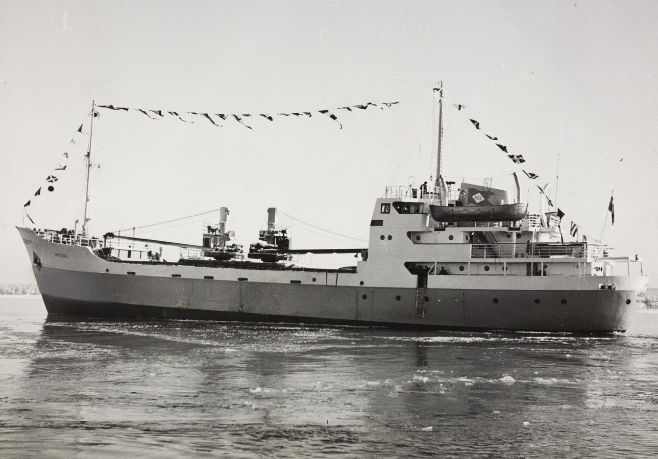 Bildet er et svart‑hvitt fotografi som viser M/S "Arendal" fra siden under jomfruturen i februar 1955. Skipet fyller nesten hele rammen og ligger på rolig vintervann med små isflak spredt i overflaten. Skipet er fotografert mot sin barbord side. Det har et langt og smalt skrog med et åpent arbeidsdekk forut og et større overbygg akter. Overbygget har flere nivåer med vinduer, rekkverk og et livbåtstativ som bærer en livbåt. Mastene står både forut og akter, forbundet med en line hvor en rekke vimpler og flagg henger som del av markeringen av jomfruturen. Midt på skipet er lastemaster og kraner tydelig synlige, plassert på hver side av en stor luke eller arbeidsflate. Kabler og taljer er strukket mellom mastene og dekksstrukturen. Skipets skrog er rent malt og uten last, noe som får det til å ligge høyt i vannet. Himmelen er lys og uten skyer, og horisonten i bakgrunnen viser et lavt landområde med vinterlige, nakne trær. Motivet formidler et maritimt og høytidsstemt øyeblikk fra skipets aller første seilas.