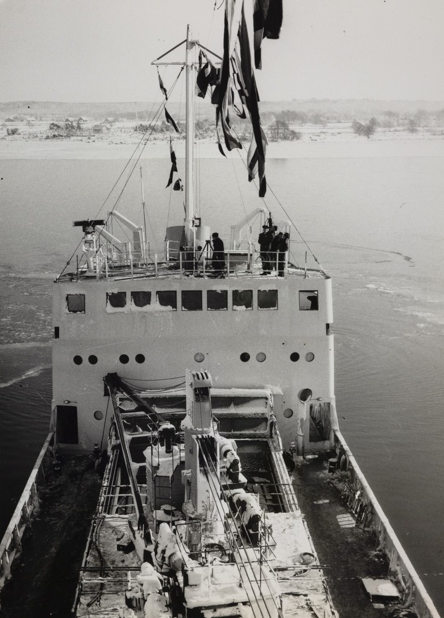 Bildet er et svart‑hvitt fotografi tatt om bord på M/S "Arendal" under jomfruturen i februar 1955. Motivet er tatt fra en høy posisjon, trolig oppe i en av mastene, og viser et fugleperspektiv ned mot skipets overbygg og fordekk. Midt i bildet ligger det store akteroverbygget, med vindusrekker og flere personer stående oppe på øverste dekk. De ser ut over akterenden og vannet bak skipet. Overbygget har en firkantet, funksjonell form typisk for 1950‑tallsskip, og flere antenner, stag og vaiere strekker seg opp fra masten og ned mot dekk. I forgrunnen sees lastedekk og arbeidsområdet på midtskipet. Her er kraner, bommer og taljer montert i en teknisk struktur. Flere personer befinner seg på dette arbeidsdekket, sittende eller stående nær lasteutstyret. Det ligger et lett lag med is eller frost på overflatene. I bakgrunnen skimtes islagt farvann og et lavt, vinterlig landskap med trær uten løv. Skipet skjærer gjennom et spor av brutt is, og vannet bak overbygget viser tydelige spor etter fart gjennom isen. Over alt dette vaier en rekke flagg og vimpler ned fra masten, som synlig markering av jomfruturen. Motivet gir en tydelig følelse av høyde, maritimt arbeid og festpreg i et vinterlig landskap.