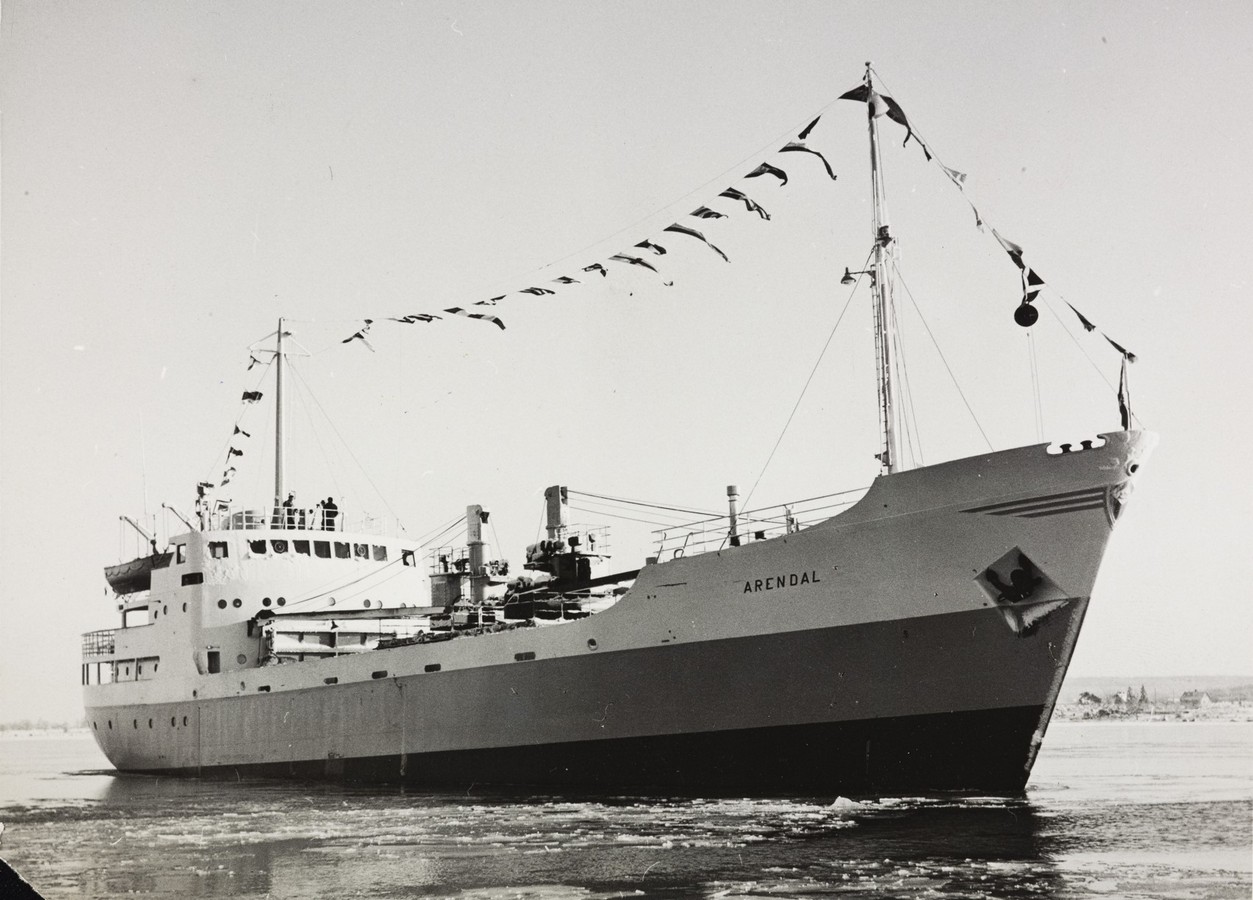 Bildet er et svart‑hvitt fotografi som viser M/S "Arendal" under sin jomfrutur i februar 1955. Skipet er avbildet fra styrbord side og fyller store deler av motivet. Det seiler gjennom et vannspeil med isflak og tynn issørpe, som sprekker opp rundt baugen. Skroget tydelig merket med navnet “ARENDAL” midt på siden. Forut står en høy mast, og fra mast til mast er det spent opp en line med en rekke flagg og vimpler som markerer den høytidelige anledningen. På fordekket er lasteutstyr synlig: master, bommer og taljer som brukes for lasting og lossing. Midtskipet har en rekke tekniske detaljer, inkludert kraner og dekksstrukturer. Overbygget akter har flere vinduer og åpne dekk med rekkverk, der flere personer står og ser ut over vannet. Himmelen er lys og klar, uten skyer. I bakgrunnen skimtes lavt, vinterlig landskap med nakne trær langs horisonten. Motivet formidler en høytidspreget og maritim stemning, der skipet viser seg fram i full størrelse på sin første seilas.