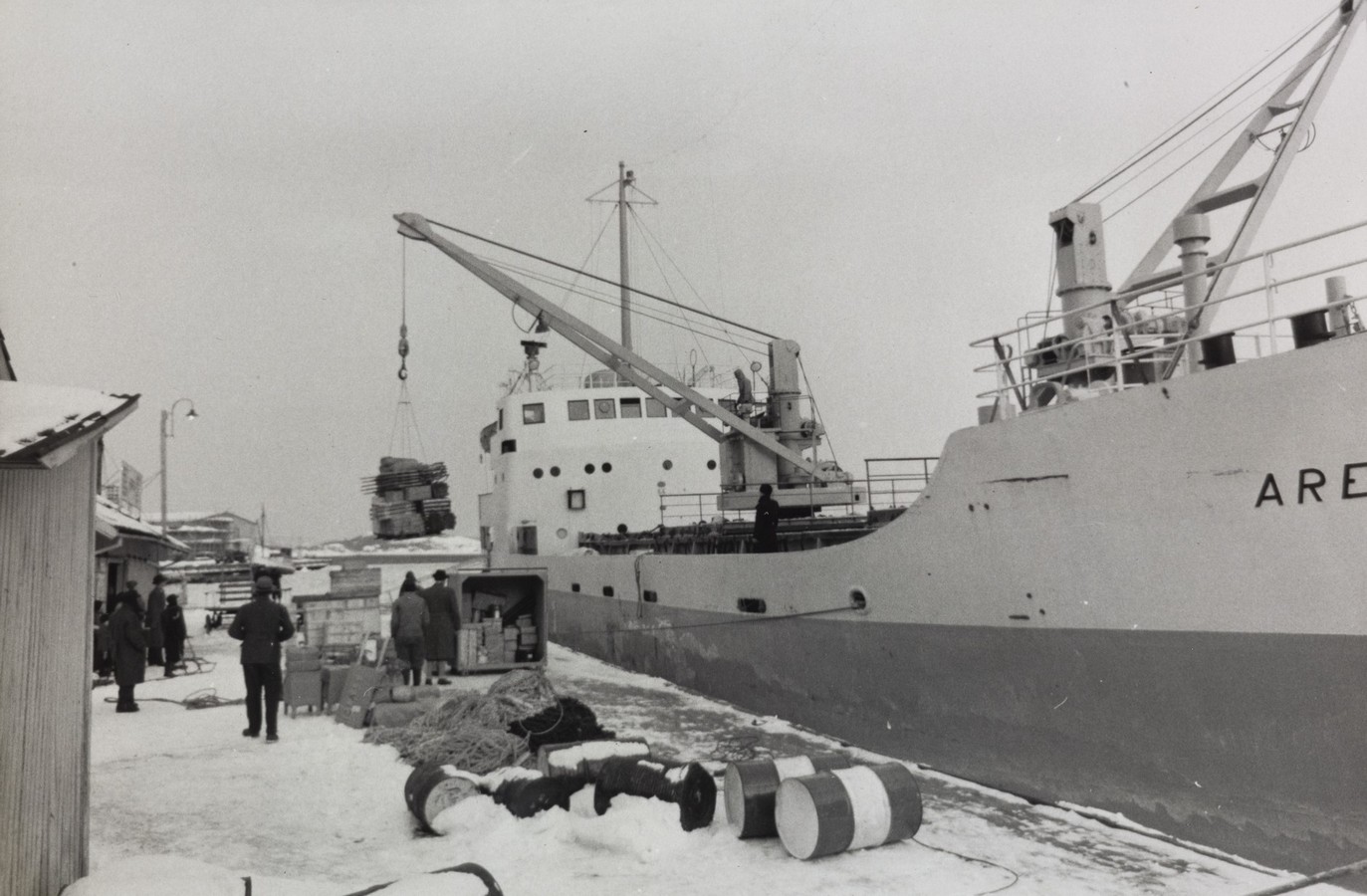 Bildet er et svart‑hvitt fotografi som viser M/S "Arendal" ved kai en vinterdag i 1955. Skipets skrog fyller høyre side av motivet, og navnet “ARENDAL” er delvis synlig på skutesiden. Dekket med lastemaster og bommer er tydelig, og en kranbom er i bruk. Midt i bildet blir en last av stablede kasser eller trekasser heist om bord eller i land med talje og krok. Lasten henger i luften, på vei mellom skipet og kaien. Ved kaifronten står flere personer i vinterklær og observerer eller deltar i arbeidet. På kaien ligger diverse utstyr og materialer spredt: store tønner, tauverk, kveiler, kasser og snødekte overflater. Til venstre står et lite skur eller havnebygg. Lenger bak skimtes flere bygninger og master fra andre fartøy. Hele scenen er preget av snø og kulde, med lyst vinterlys og et stille havneområde. Motivet gir et troverdig inntrykk av daglig arbeid i havnen, der M/S "Arendal" blir håndtert på tradisjonelt vis i et vinterlig miljø.