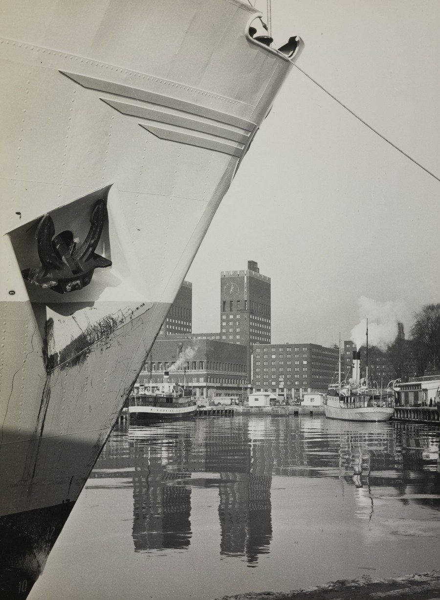 Bildet er et svart‑hvitt fotografi som viser M/S "Arendal" liggende til kai i Oslo en vinterdag i 1955. I forgrunnen fyller baugen av skipet venstre side av motivet. Ankeret er festet i skipssiden, og mørke striper fra vann eller frost renner nedover skroget. En fortøyningstrosse går oppover fra baugen og ut av bildet. Vannet er rolig og gjenspeiler både skipet og bygningene i havnebassenget. Lenger bak i bildet, på motsatt side av fjorden, står Oslo rådhus med sine to karakteristiske tårn. Rundt kaianlegget ligger flere havnebygninger og mindre fartøy, blant annet småbåter og arbeidsbåter. Til høyre i motivet skimtes en vinterlig skråning med trær uten løv og enkelte bygninger langs kanten. Lett damp eller røyk stiger fra et punkt i bakgrunnen, og det er tydelig at temperaturen er lav, med snø liggende flere steder. Motivet gir et rolig og vinterlig inntrykk av Oslo havn på 1950‑tallet, med M/S "Arendal" dominerende i forgrunnen og rådhuset som et klart landemerke i bakgrunnen.