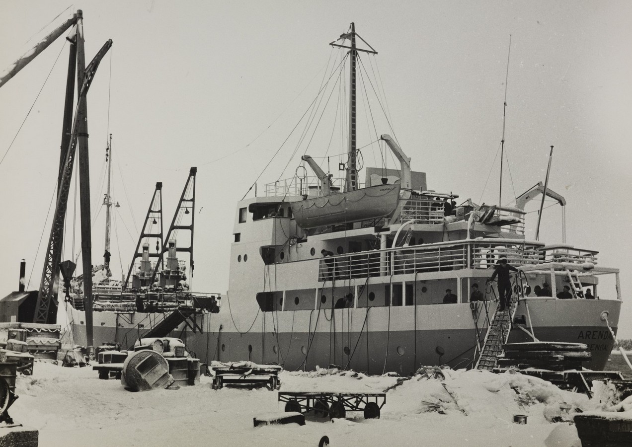 Bildet er et svart‑hvitt fotografi som viser M/S "Arendal" liggende til kai om vinteren, i et område med industriell aktivitet. Skipet fyller store deler av motivet, sett fra babord side og litt akterover. Overbygget har flere nivåer med vinduer og rekkverk, og en livbåt ligger plassert i daviter på øvre dekk. Master, antenner og taljer strekker seg opp mot den lyse vinterhimmelen. Langs kaien står ulike maskiner og utstyr, blant annet en stor kran med lang arm på venstre side av bildet. Snø dekker både bakken og mange av gjenstandene som ligger rundt — tønner, planker, stålrør, kasser og annet havnemateriell. I forgrunnen, til høyre, er en person på en skrå stige som står lent mot skipssiden. Personen arbeider med noe på dekk eller rekkverk. Flere tau, vaiere og kabler henger ned langs skutesiden og er festet i kaien. Lenger bak mot venstre sees andre fartøy og kraner, som antyder et travelt havne- eller verftsområde. Hele scenen fremstår kald og snødekt, med tydelig vinterpreg og rolig aktivitet rundt et fortøyd skip.