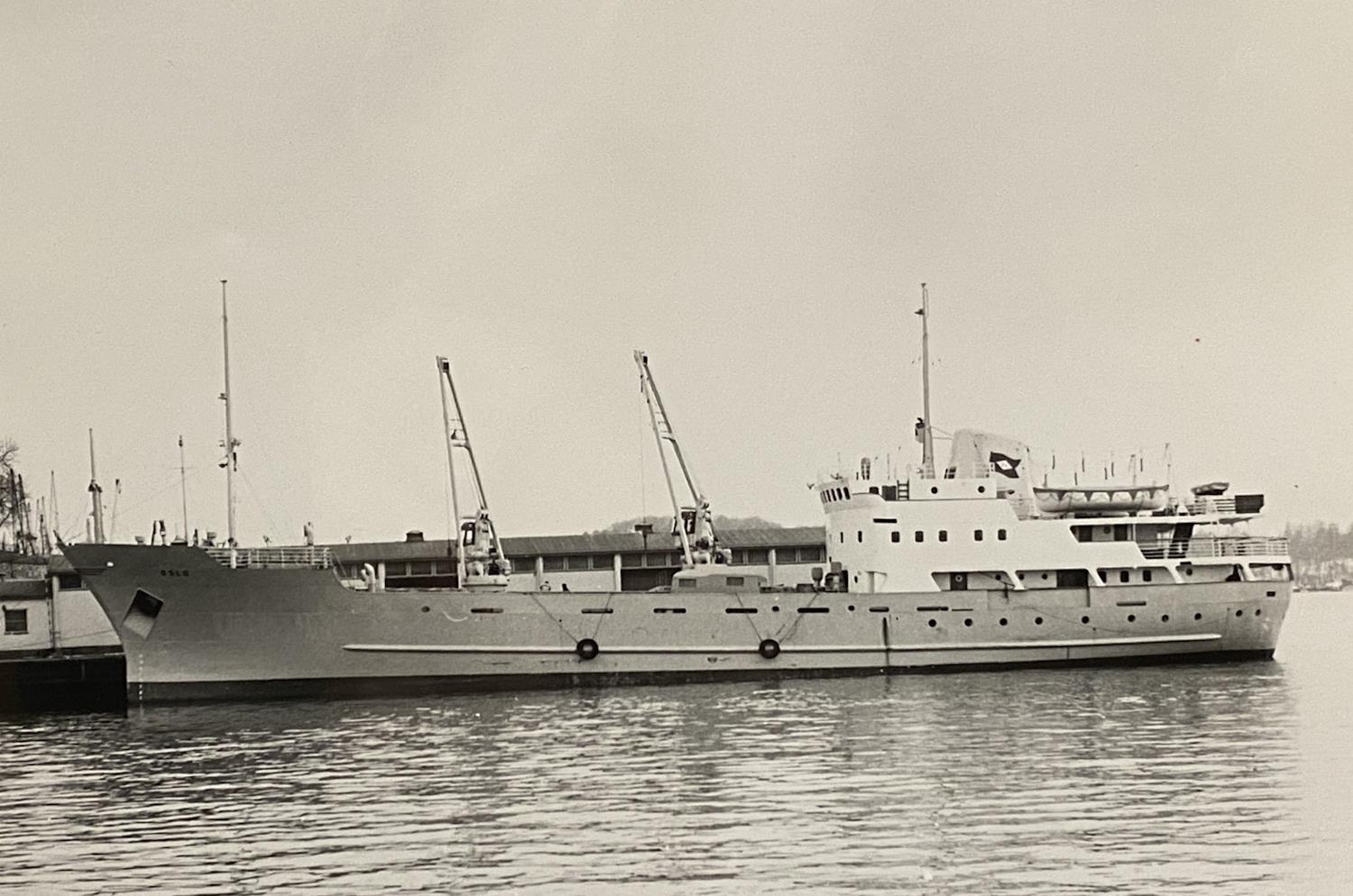 Et sort‑hvitt‑fotografi viser et større lasteskip som ligger ved kai. Skipet har en lang, jevn skrogside og to lastekraner plassert midtskips. Akterdelen har en høy overbygning med flere etasjer, rekkverk og små vinduer som følger skipets linjer. Vannet er stille og gir et svakt speilbilde av skroget. I bakgrunnen skimtes land og lave bygninger langs havneområdet under en lys, overskyet himmel. Atmosfæren i bildet er rolig og tydelig preget av tidsepoken det er tatt i.