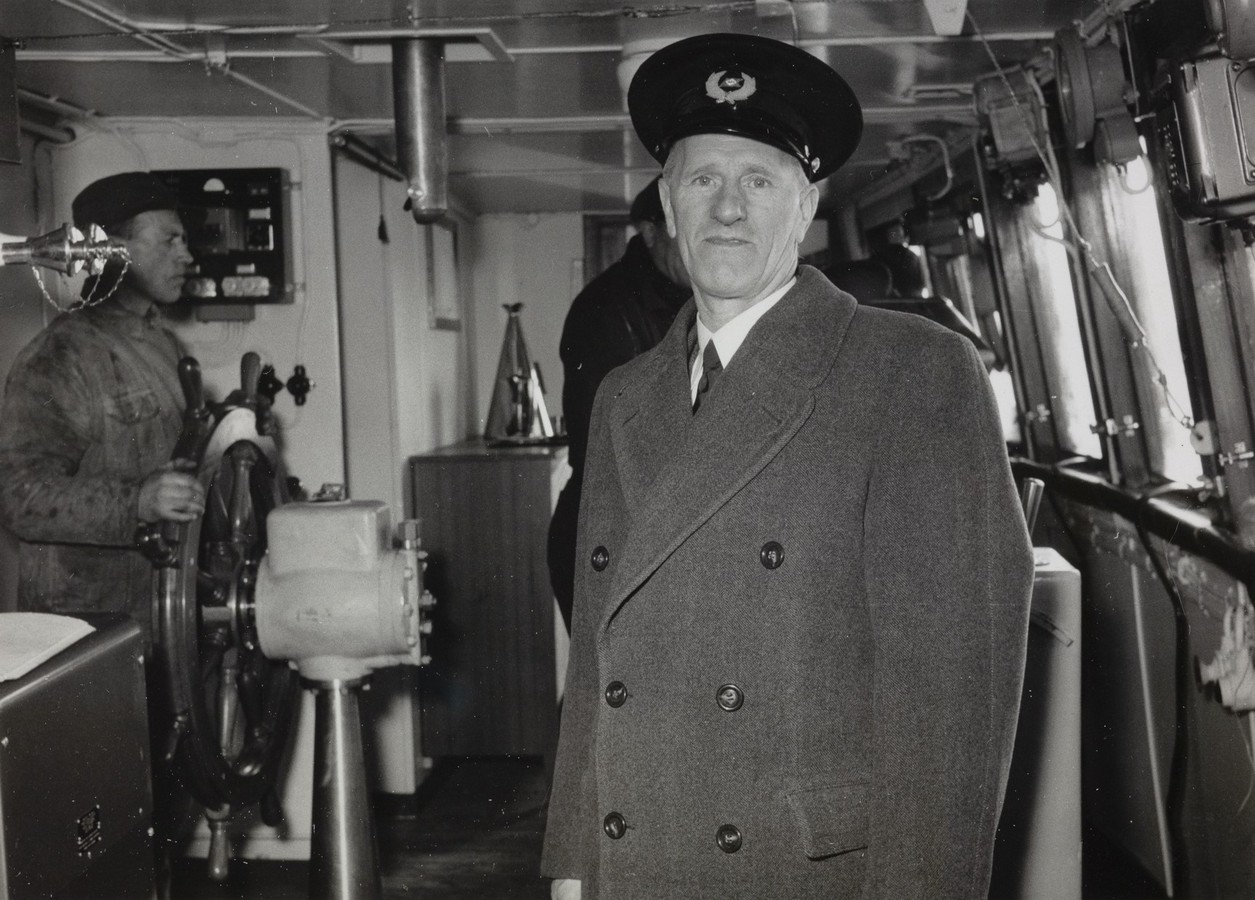 Bildet er et svart‑hvitt fotografi tatt inne på broa om bord i M/S "Arendal" i 1955. Motivet viser et arbeidsmiljø fullt av navigasjons- og styringsutstyr fra perioden. I forgrunnen står en person i uniform. Vedkommende har på seg en lang, mørk frakk med doble knapper og en uniformslue med emblem. Personen står vendt mot kamera, plassert litt til høyre i bildet. Bak vedkommende, lenger inn på broa, står en annen person ved skipets ratt. Rattet er stort og av tradisjonell type, montert på en kraftig metallkonsoll. Like ved finnes andre mekaniske instrumenter og kontrollpaneler typiske for styring av skip på 1950‑tallet. Langs høyre side av bildet er en rekke vinduer som slipper inn lys og gir utsikt til skipets utside. Under vinduene løper en smal hylle med ulike små gjenstander og utstyr. På venstre side av rommet henger tekniske paneler, brytere og kabler. Taket har synlige rør, ventiler og konstruksjonselementer som understreker broas funksjonelle og tekniske karakter. Stemningen i bildet er profesjonell og maritim, med fokus på arbeidssituasjonen om bord.