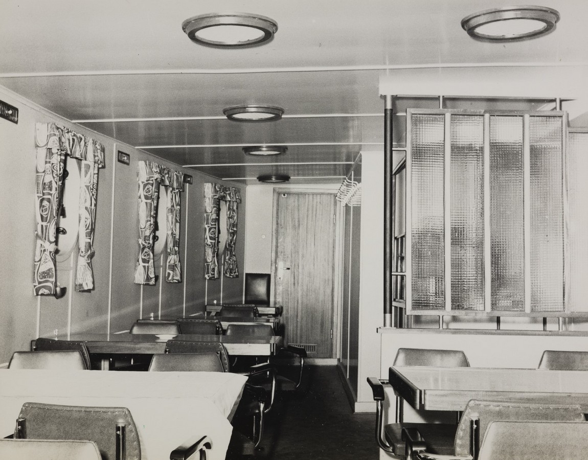 Bildet er et svart‑hvitt fotografi fra spisesalen om bord i M/S "Arendal" i 1955. Rommet er avlangt og innredet med flere bord og stoler plassert i rekker langs venstre vegg. Bordene er rektangulære, og noen av dem har duk. Stolene har enkle metallrammer og polstrede seter og rygger. Langs venstre side av rommet er det flere runde skrogvinduer med brede metallrammer. Disse er dekket med mønstrede gardiner i et karakteristisk 1950‑tallsdesign med store figurer og sterke kontraster. Veggene er lyse og glatte, med enkel panelinndeling. I taket henger runde, innfelte lamper som gir et jevnt lys. Midt i bildet, mot høyre, står en vegg eller romdeler med dekorpanel bestående av rutete glassplater i en metallramme. Denne avskjermingen skaper et skille mellom spisesalen og et annet rom eller område. Lenger bak i rommet, midt på bakveggen, står en enkel dør i tre med en liten jakkehenger over. Bakdøren skimtes et skap eller en liten benk. Interiøret fremstår som funksjonelt, ryddig og typisk for en maritim spisesal på 1950‑tallet, med klare linjer og praktiske løsninger.