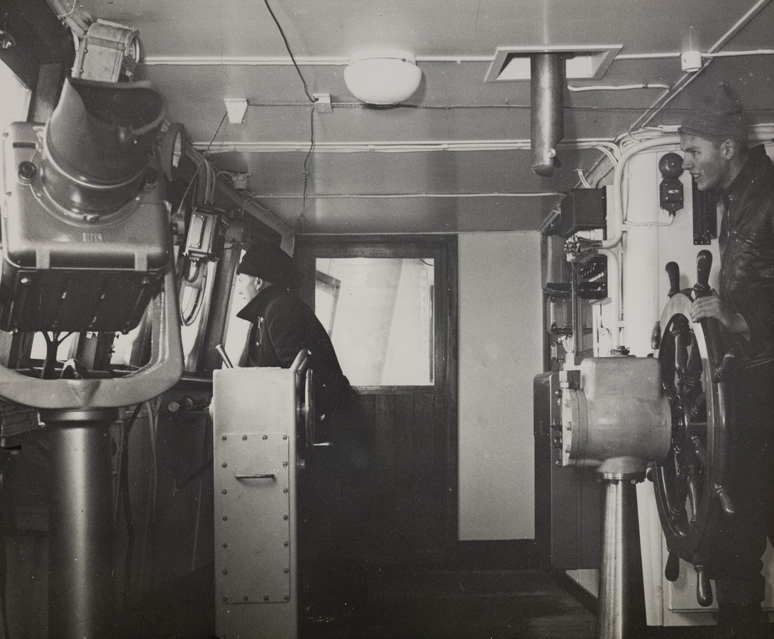 Bildet er et svart‑hvitt fotografi som viser interiøret på broa om bord i M/S "Arendal" i 1955. Motivet er tatt fra fra barbord side og i enden er en dør til styrbord broving. Til venstre i bildet står et stort navigasjonsinstrument montert på en solid metallstang. Instrumentet har et sikterør eller kikkertlignende toppdel. Lenger forover står en person ved et navigasjonsbord og ser ut gjennom brovinduene. Personen bærer uniform eller mørke klær. Til høyre i bildet står en person ved skipets ratt. Rattet er av klassisk type med flere håndtak og er festet til en kraftig styrekonsoll i metall. Rundt konsollen finnes brytere, indikatorer og kabling som hører til styringskontrollene. Taket er lavt og preget av synlige rør, ledninger og ventilasjonskanaler. Midt i taket henger en rund taklampe. Rommet har et funksjonelt preg med lyse vegger og vinduer som vender ut mot sjøen. Motivets uttrykk er teknisk og maritimt, med fokus på arbeidssituasjonen på broa under skipets drift.