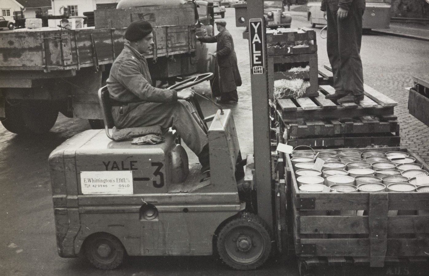 Bildet er et svart‑hvitt fotografi som viser en truck i arbeid på en kai i 1955. Trucken, av merket YALE, står i midten av motivet og løfter en stor trekasse fylt med tønner eller metallbeholdere. Kassen er solid bygget, med tverrbjelker og remmer som holder innholdet på plass. Føreren sitter på truckens venstre side, kledd i tykke arbeidsklær som passer et kaldt, utendørs havnemiljø. På truckens side er den tydelig merket med tallet 3 og en etikett fra firmaet E. Whittington Eftf., som kan tyde på utleie eller tilhørighet. Foran trucken ligger flere trepaller med lignende beholdere, og en person står på en av pallene for å assistere med arbeidet. Litt lenger bak står en annen person ved en lastebil og håndterer varer eller snakker med sjåføren. Rundt dem er det flere kjøretøy, trekasser og gods, typisk for en travel laste- og losseplass. Underlaget består av brostein eller asfalt, og det virker å være kjølig vær, men uten synlig snø. Motivet formidler et aktivt og funksjonelt havnemiljø på 1950‑tallet, der trucker og mannskap arbeider tett sammen for å håndtere lasten.