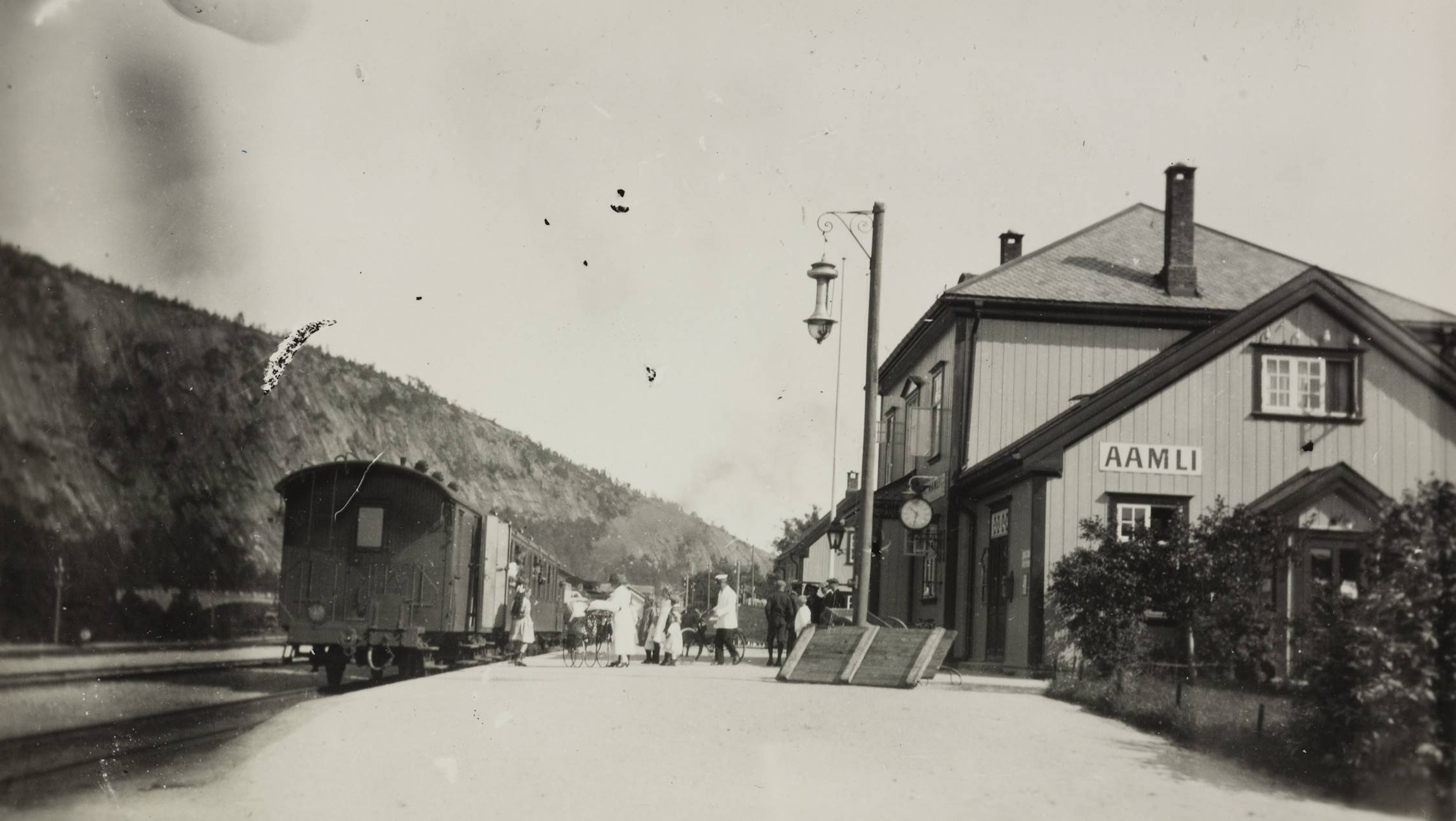 Åmli stasjon. Bildet viser et tog som står på jernbanesporet ved siden av en perrong. Til høyre står en toetasjes trebygning med skiltet «AAMLI». Foran bygget står en gatelykt på en høy metallstolpe. På perrongen står en gruppe mennesker, blant annet barn og voksne, samlet i nærheten av toget. I bakgrunnen sees skogkledde åssider og blå himmel. Bildet har et rolig sommerpreg.