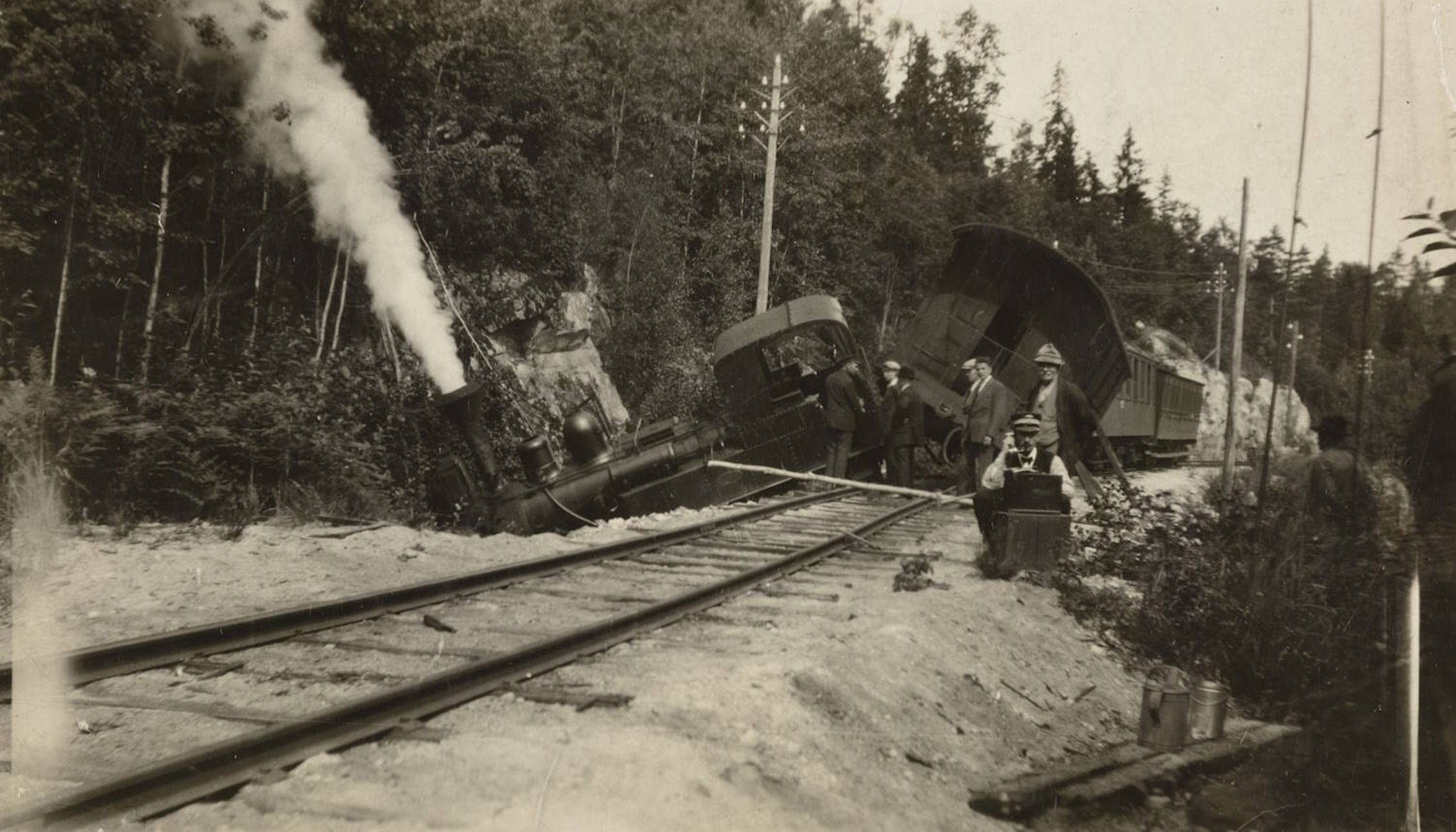 Avsporingen ved Bøylefoss 05.08.1928. Bildet viser et tog som har sporet av i et skogsområde. Lokomotivet ligger på skrå ned i grøftekanten ved siden av skinnene, og det kommer hvit damp eller røyk opp fra det. Flere togvogner bak lokomotivet ligger også veltet og delvis skadet. Rundt toget står flere menn i hatt og arbeidsklær og undersøker skadene. I forgrunnen ser man togskinnene som går i en svak kurve, og i bakgrunnen tett skog og telefonstolper langs traseen. Stemningen er rolig og dokumenterende.