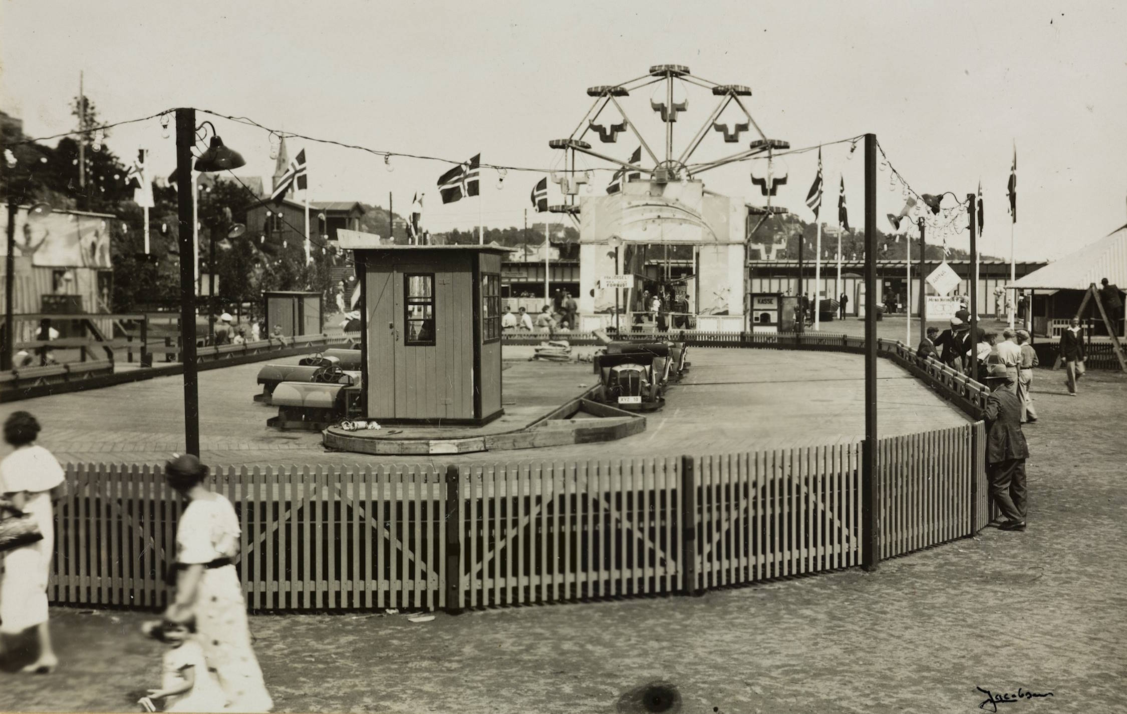 Bilbanen i fornøyelsesavdelingen på Idrettsplassen under varemessen i Arendal i 1936. Foto: Jacobsen. Bildet viser en utendørs fornøyelsesattraksjon på et messeområde. I forgrunnen står en oval innhegning omgitt av et lavt gjerde med tettstilte trestakitter. Innenfor gjerdet ligger en kjørebane for små elektriske biler. Banen er flat og har en liten trehytte plassert omtrent i midten. Rundt hytta står flere små støtfangerbiler parkert, og noen står delvis ut på banen. Over banen henger en rekke flagg i en snor — blant annet landsflagg — festet mellom høye stolper. Det henger også flere små lamper fra samme snor, som lyspærer i en dekorativ belysningsrekke. I bakgrunnen står et pariserhjul. På begge sider av banen er det mange mennesker som går rundt eller står og ser utover området. De er kledd i 1930‑tallsmote — kvinner med kjoler og hatter, menn med dresser og hatter. Lenger bak til høyre står et stort telt med spisst tak. På venstre side av bildet skimtes flere boder og bygg som hører til fornøyelsesavdelingen. Stedet har tydelig preg av en travel og livlig messeplass fra mellomkrigstiden.