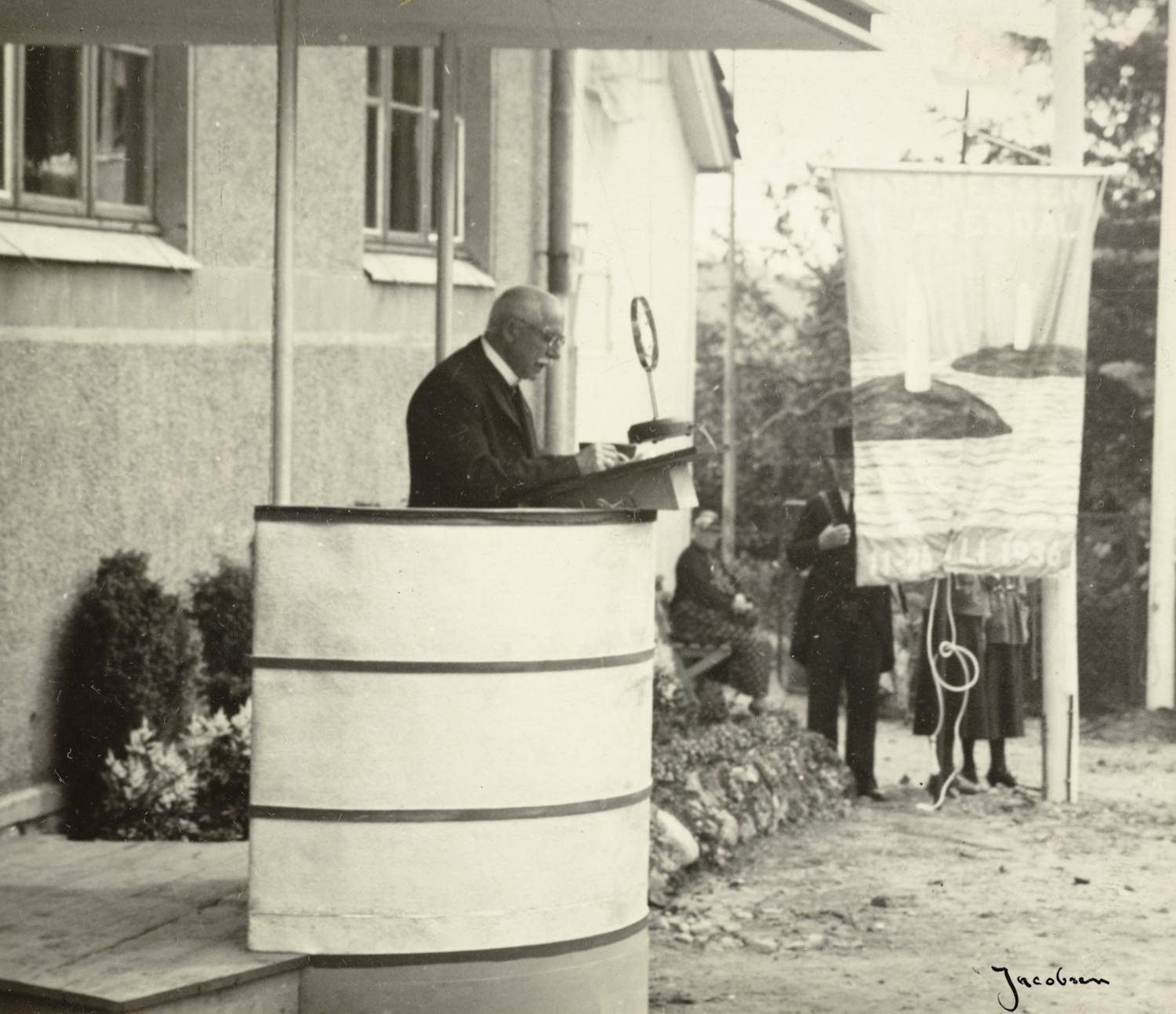 Bryggerieier Wilhelm Christiansen holder tale ved åpningen av varemessen i Arendal 11.07.1936. Foto: Jacobsen. En mann står ved en høy, sylinderformet talerstol foran skolebyggets murvegg. Talerstolen har horisontale mørke striper og står under et lite overbygg. Ved siden av står en vimpel eller fane med bilde av et landskap og tekst nederst.