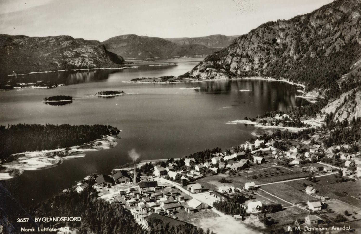 Byglandsfjord rundt 1950. Bildet viser et svart‑hvitt luftfoto av Byglandsfjord. Nederst i bildet ligger et lite tettsted med lave bygninger, spredte hus og rette veier. Røyk stiger opp fra en fabrikkpipe i den nederste delen av bildet. Landskapet rundt består av skogkledde områder og åpne plener. I midten av bildet ligger fjorden med rolig vann og flere små øyer. På begge sider av fjorden stiger bratte fjellsider med skog. I bakgrunnen ses fjorden som strekker seg innover mellom fjellene. Nederst til venstre står trykt teksten «3657 Byglandsfjord» og «Norsk Luftfoto». Nederst til høyre står navnet på fotografen og byen vedkommende var basert i.
