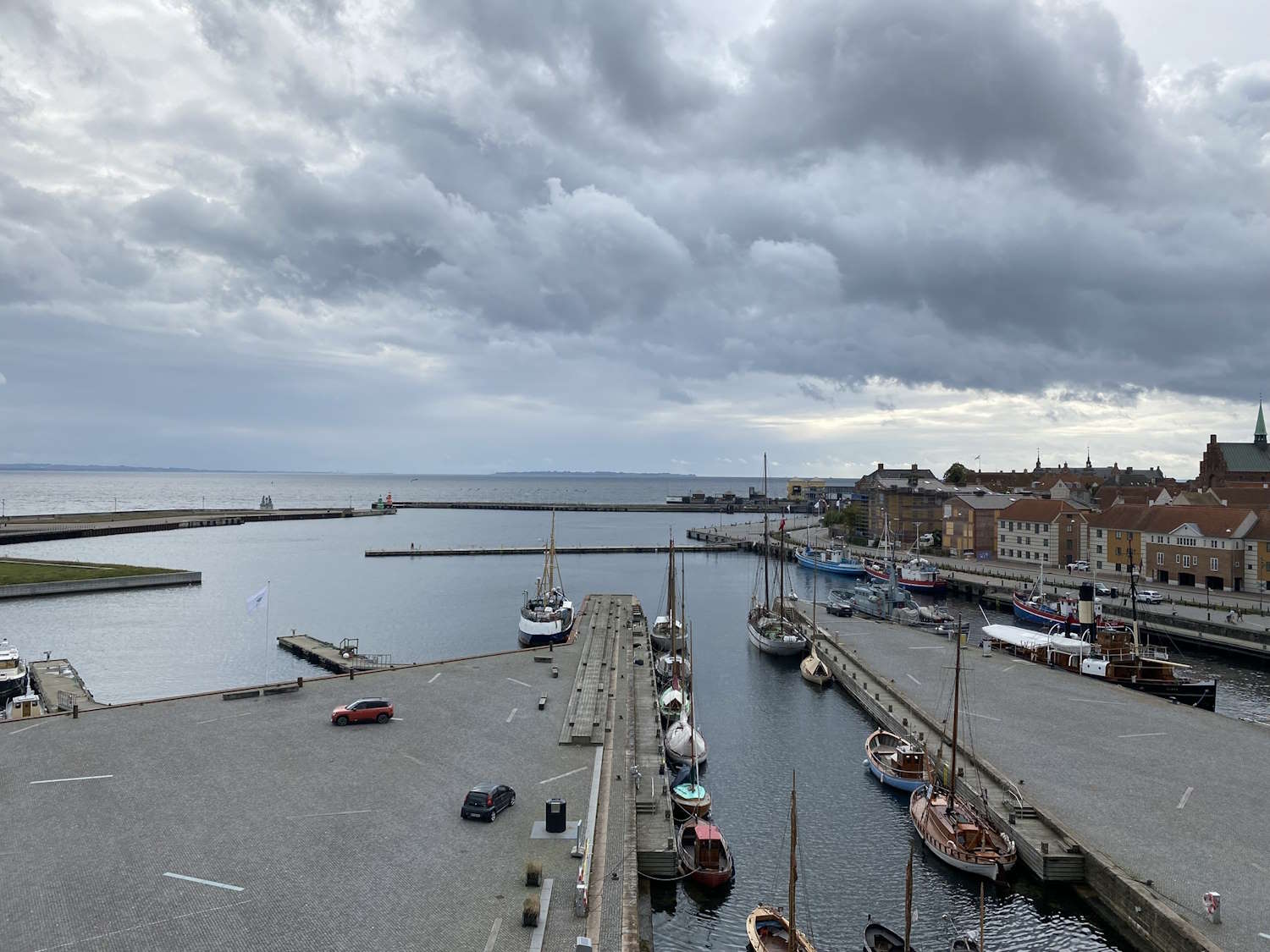 Fotografiet viser deler av havnen i Helsingør sett fra et forhøyet ståsted. I forgrunnen er det et stort, åpent kaiområde belagt med grå stein. På plassen står noen få biler parkert. Midt i bildet går en bred molo utover mot sjøen, med flere fortøyde seilbåter langs begge sider. Mastene stikker høyt opp og danner en vertikal kontrast til det åpne landskapet. Til høyre i bildet ligger en annen kaiarm med flere båter langs kanten, blant dem flere trebåter. Bak denne kaien ses en rekke eldre, mursteinfargede bygninger langs vannkanten, med et kirketårn og andre spisse tak som stikker opp i bysilhuetten. Ute i havnebassenget er vannet rolig. Lengst ute sees lave moloer som rammer inn innseilingen. I horisonten skimtes svenskekysten på den andre siden av sundet. Over hele motivet ligger en dramatisk himmel med tunge, grå skyer som dekker store deler av himmelen. Dette gir et høstlig og litt mørkt preg til scenen.