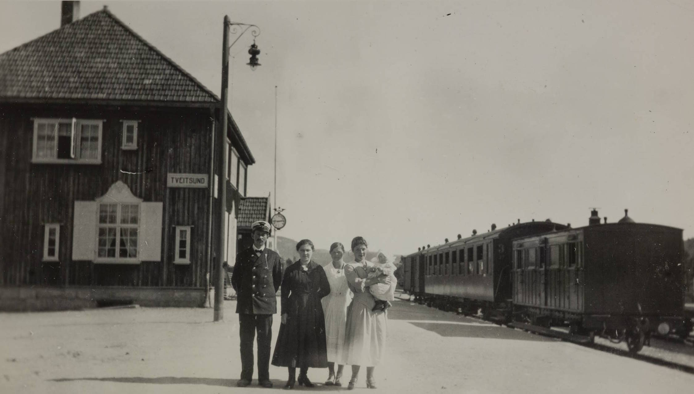 Familien Stien utenfor Tveitsund stasjon 1920. Foran stasjonsbygningen står fem personer oppstilt for fotografen. Tre voksne kvinner, én mann i uniform og én kvinne som holder et lite barn i armene. Stasjonsbygningen til venstre har skiltet «TVEITSUND». Til høyre står et tog med flere mørke passasjervogner langs perrongen. En gatelykt på stolpe står mellom bygningen og toget. Området er åpent og lyst, med klar himmel.