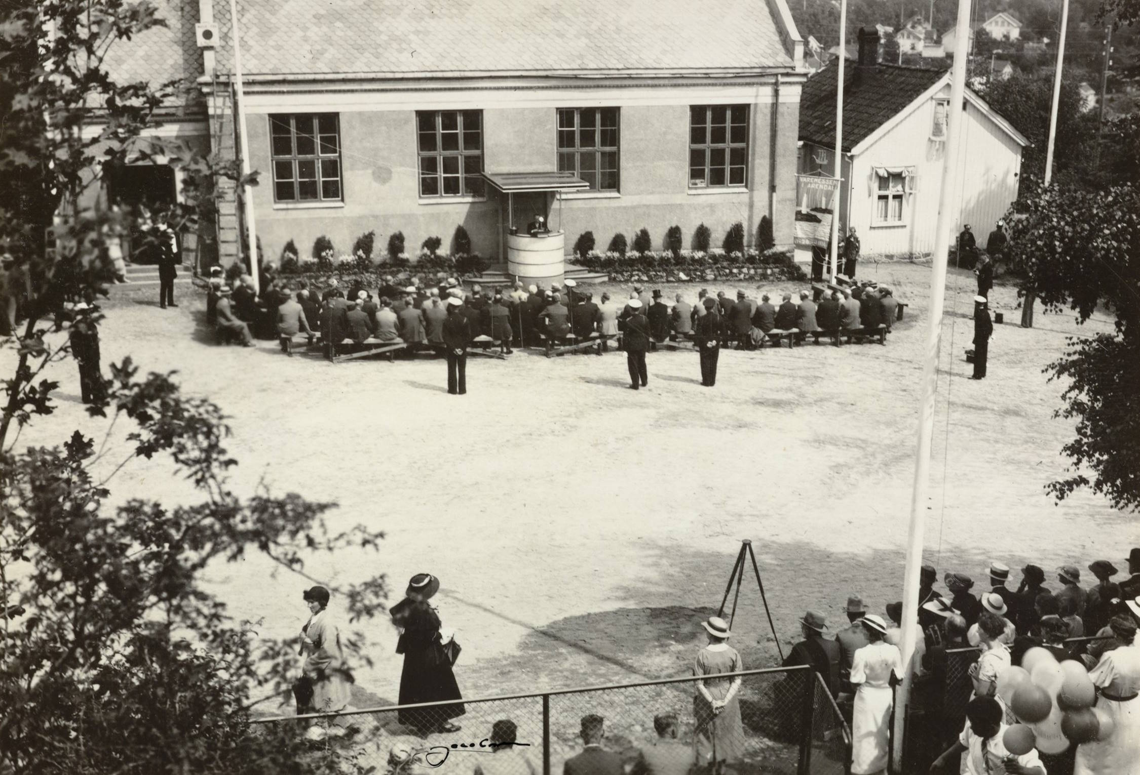 Festplassen utenfor gymnastikksalen ved Barbu skole under åpningen av varemessen i Arendal 11.07.1936. Foto: Jacobsen. Bildet viser en større samling mennesker på en åpen plass foran en mursteinsbygning med skrått tak. Bygningen har tre store vinduer og en liten talerstol eller avsats midt på veggen. En person står bak talerstolen og ser ut til å holde en tale. Rett foran talerstolen sitter flere rekker med mennesker på benker, alle vendt mot taleren. Bak disse står og sitter flere spredte grupper av mennesker, de fleste kledd i tidstypiske 1930-tallsklær — menn i mørke dresser og hatter, kvinner i kjoler og hatter. Plassen er åpen og lys, trolig gruslagt. Til høyre i forgrunnen står en gruppe mennesker bak et gjerde, som om de følger arrangementet på avstand. Et stort flagg henger fra en flaggstang ved høyre bildekant. Noen trær og busker rammer inn motivet langs venstre og høyre side. Bak bygningen skimtes et mindre trehus og bebyggelse oppover en skråning. Stemningen er høytidelig og fokusert, som ved en offisiell åpning eller markering.