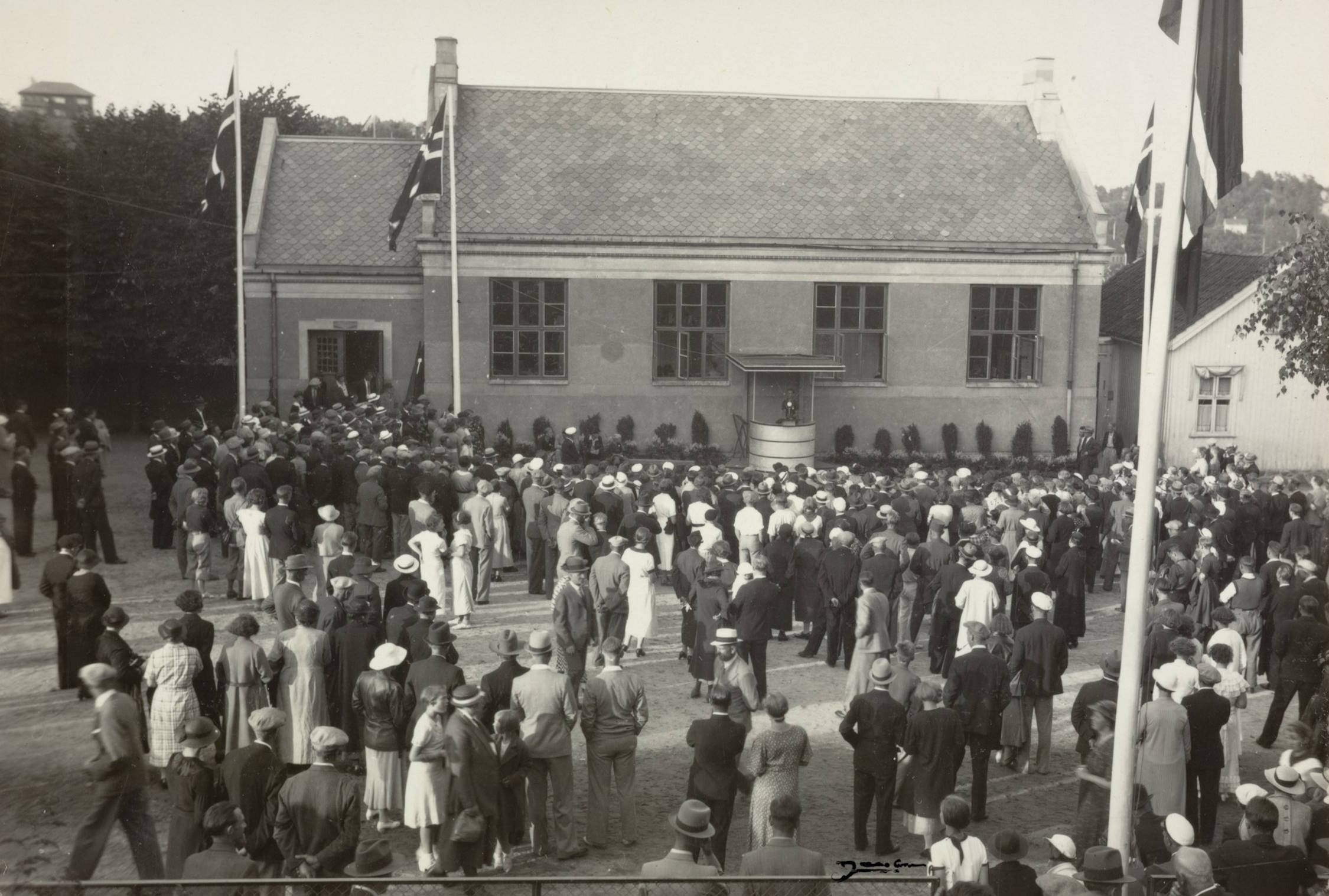 Festplassen utenfor gymnastikksalen ved Barbu skole under varemessen i Arendal 11.07.1936. Foto: Jacobsen. Bildet viser en stor folkemengde samlet foran en mursteinsbygning med skrått skifertak. Bygningen har tre høye vinduer på langveggen og en liten, firkantet talerstol eller veranda midt på fasaden, der en person står og holder tale. Foran bygningen står flaggstenger med norske flagg heist. I forgrunnen og midten av bildet står det tett med mennesker, mange kledd i mørke frakker, hatter og kåper – typiske 1930-tallsklær. Noen kvinner har lyse kjoler og hatter, mens mennene i hovedsak har mørke dresser og hodeplagg. Folk står vendt mot taleren, og det virker å være et offentlig arrangement eller høytidelig markering. Til høyre i bildet står en mindre trebygning, og i bakgrunnen sees trær og ei bakkeskråning. Flagget til høyre er plassert i nær forgrunn og fyller bildekanten. Terrenget foran bygningen består av en åpen plass, trolig gruslagt. Stemningen virker høytidelig og preget av forventning eller markering, med en stor, ordnet folkemengde vendt mot taleren.